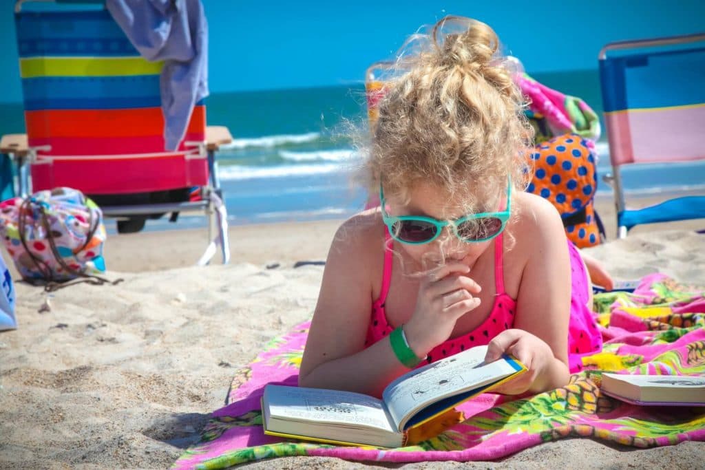 A girl wearing sunglasses reading laying down on a beach towel reading a book