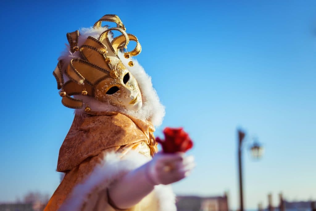 A person wearing a white and gold Venetian mask, which looks like a porcelain doll