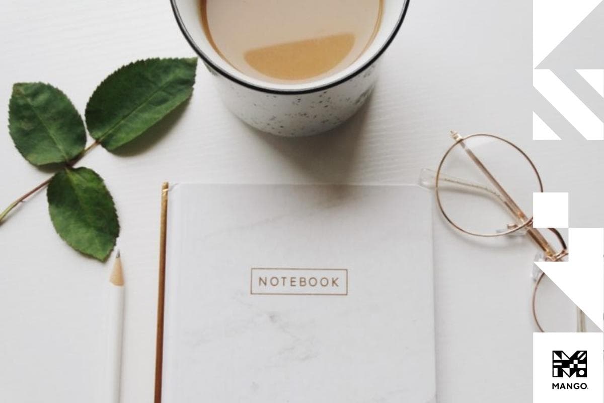 A white notebook on a table sitting next to a cup of coffee, and a pair of glasses