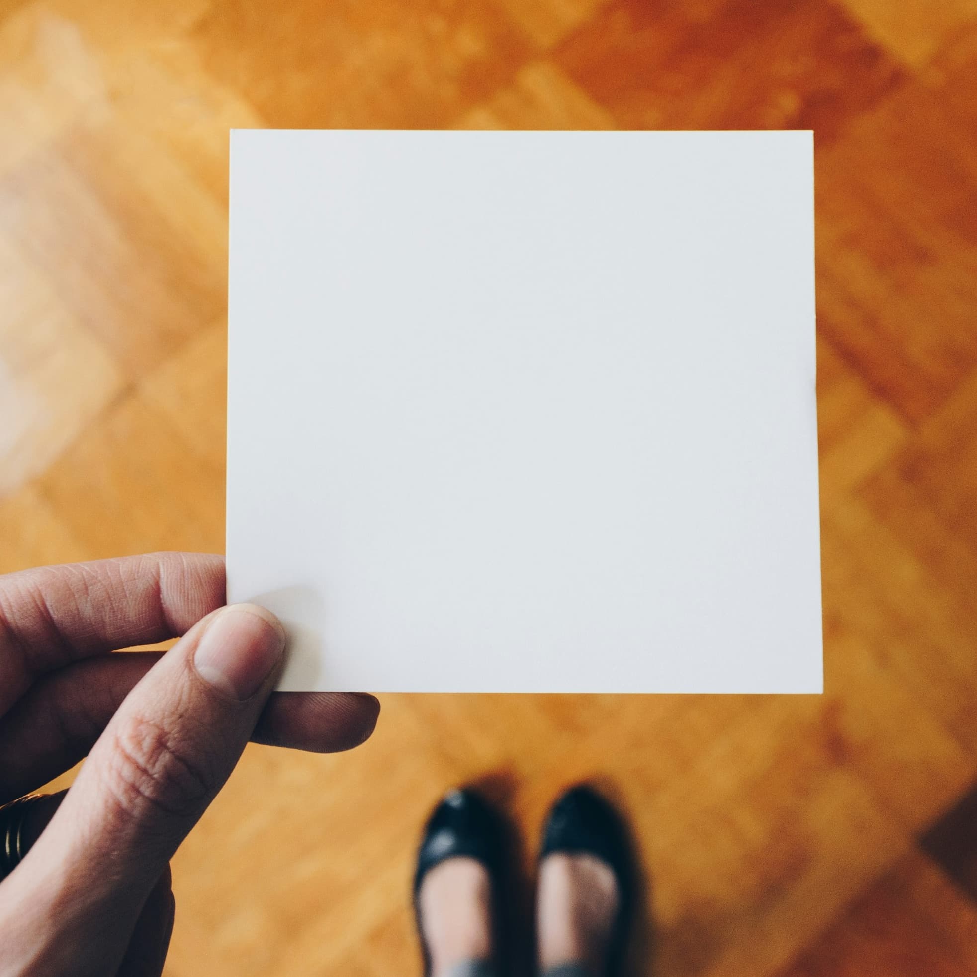 A photo looking down at a hand holding a small square of white paper. Below the paper we can see a parquet floor and the shoes of the person holding the paper.