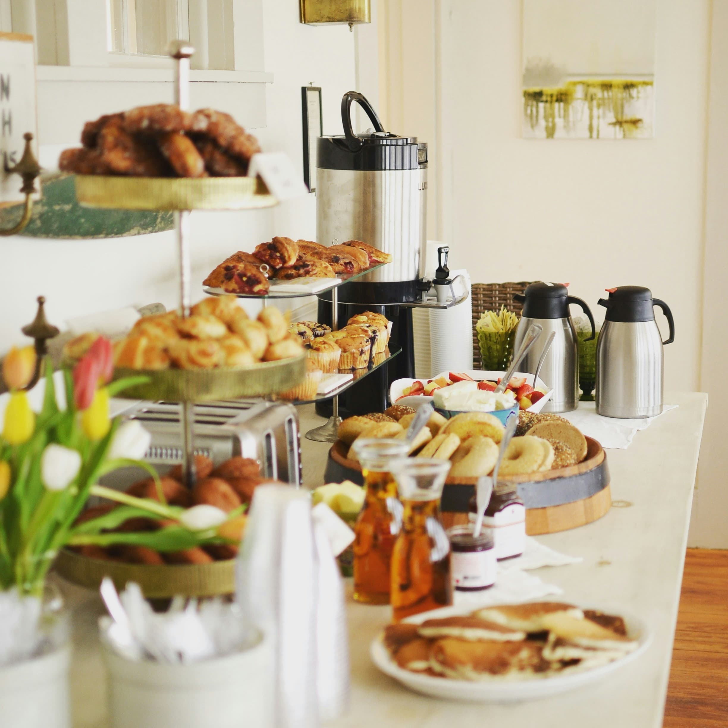 A breakfast buffet. There is a good spread of bagels and pastries, some paper plates, coffee and tea, and a vase of tulips on a white table.