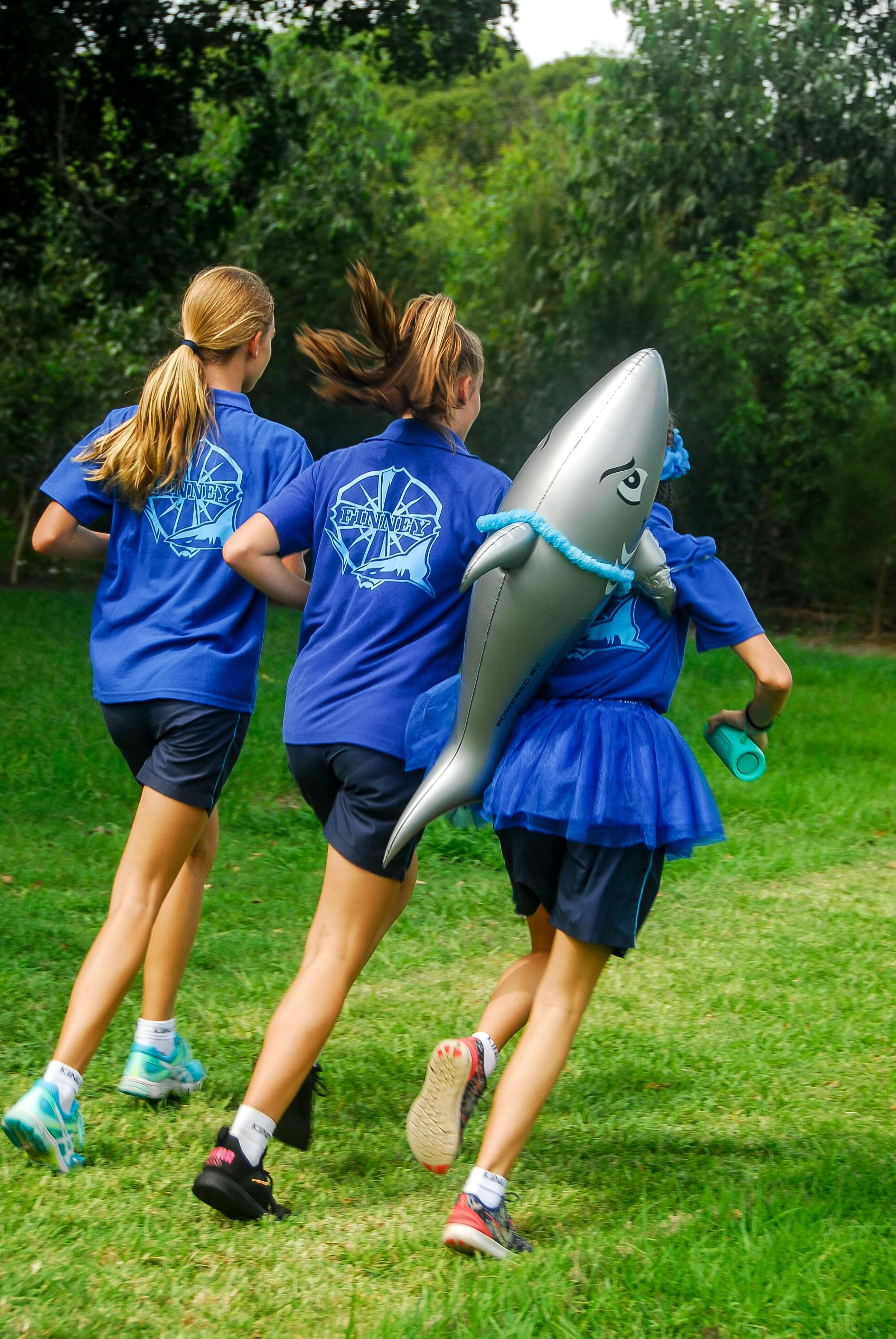A photo of three young women taken from behind. They are wearing matching outfits - blue t-shirts with a shark logo and dark running shorts - and are running away from the camera. The girl on the right has an inflatable shark strapped to her back.