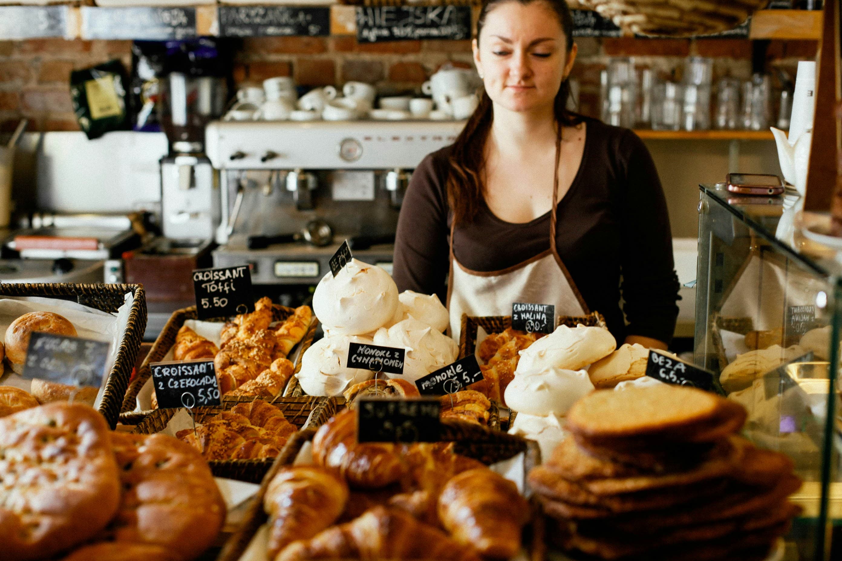 A bakery employee behind the counter. There are all sorts of delicious-looking pastries in front of her.