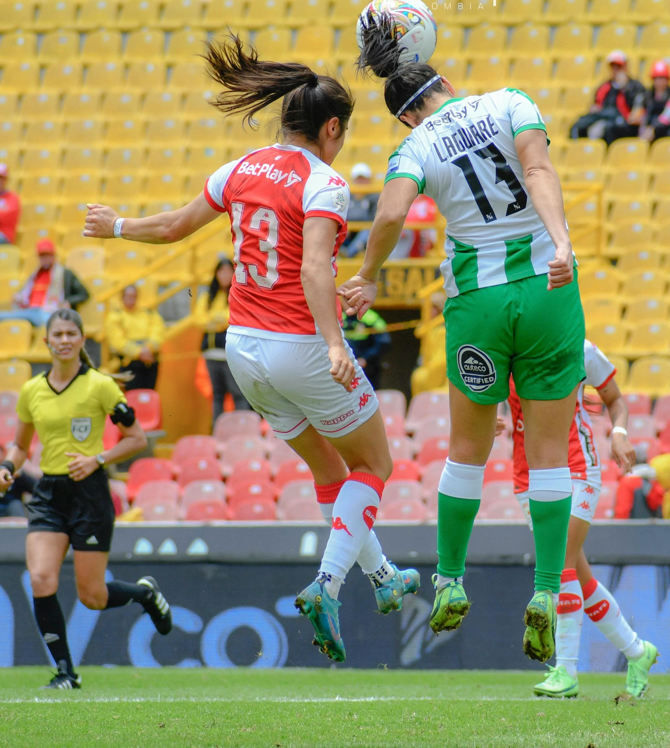 A photo of two women with long hair playing soccer. One is in a red uniform, the other in a green uniform and the photo has been taken from behind, while they are both jumping into the air trying to head the ball.