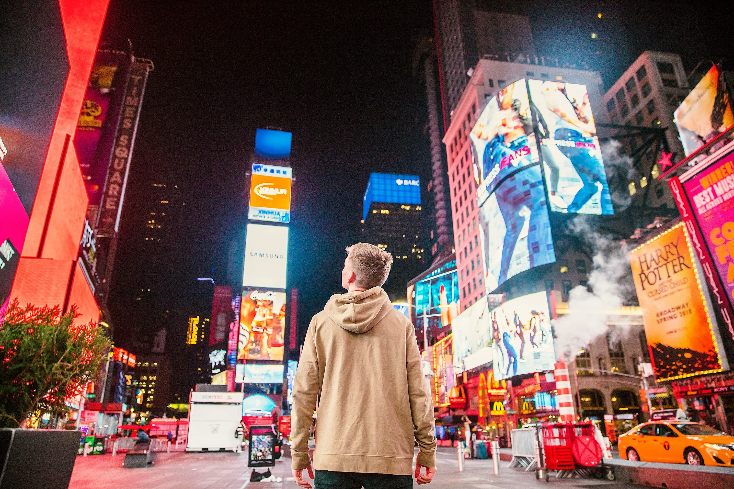 A photo of a teenage boy in Times Square, New York. The photo is taken from behind and he is looking up at all the billboards.