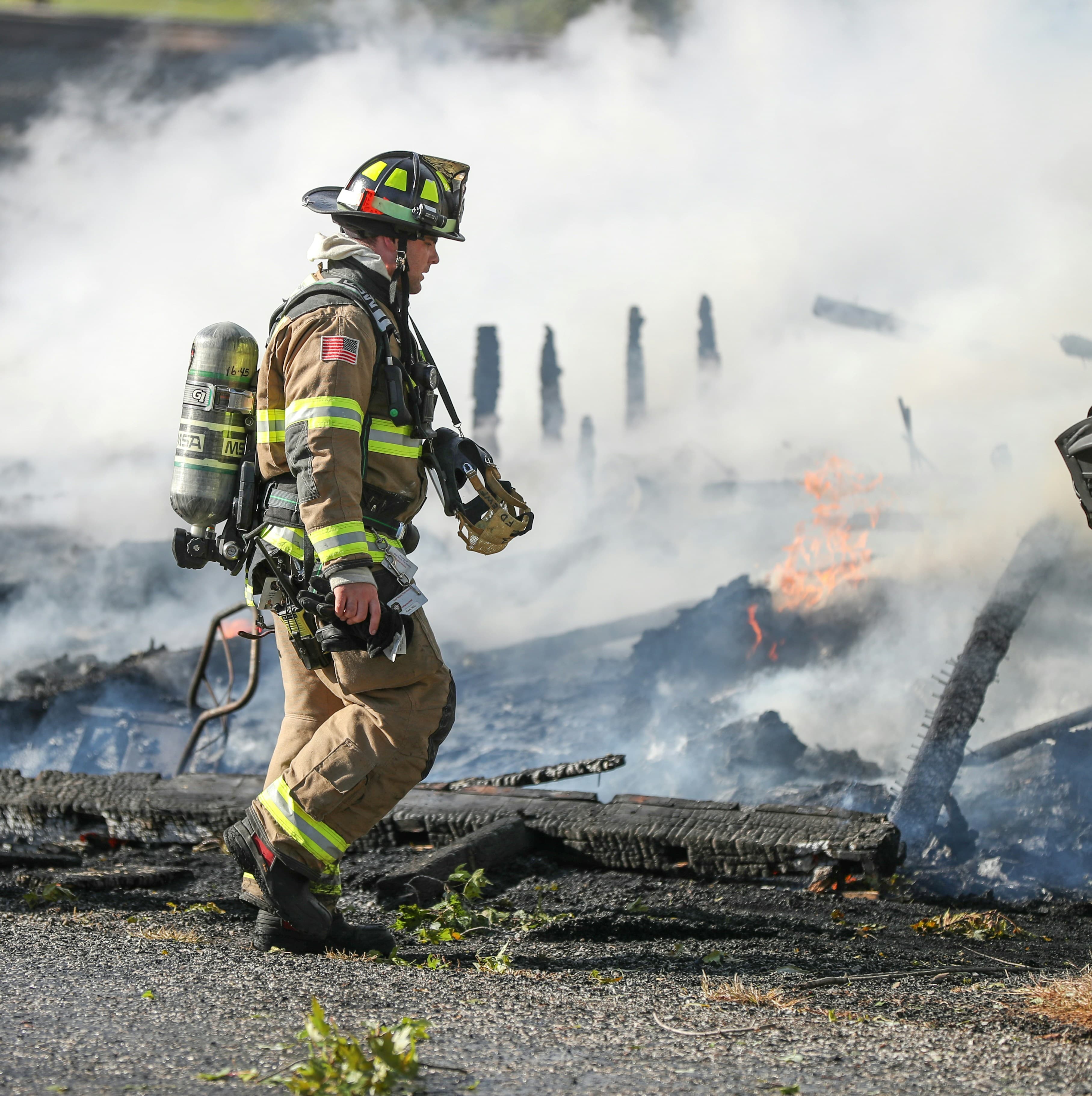 A white fireman in full brown and yellow work gear walks through the smoking remains of a burned building.