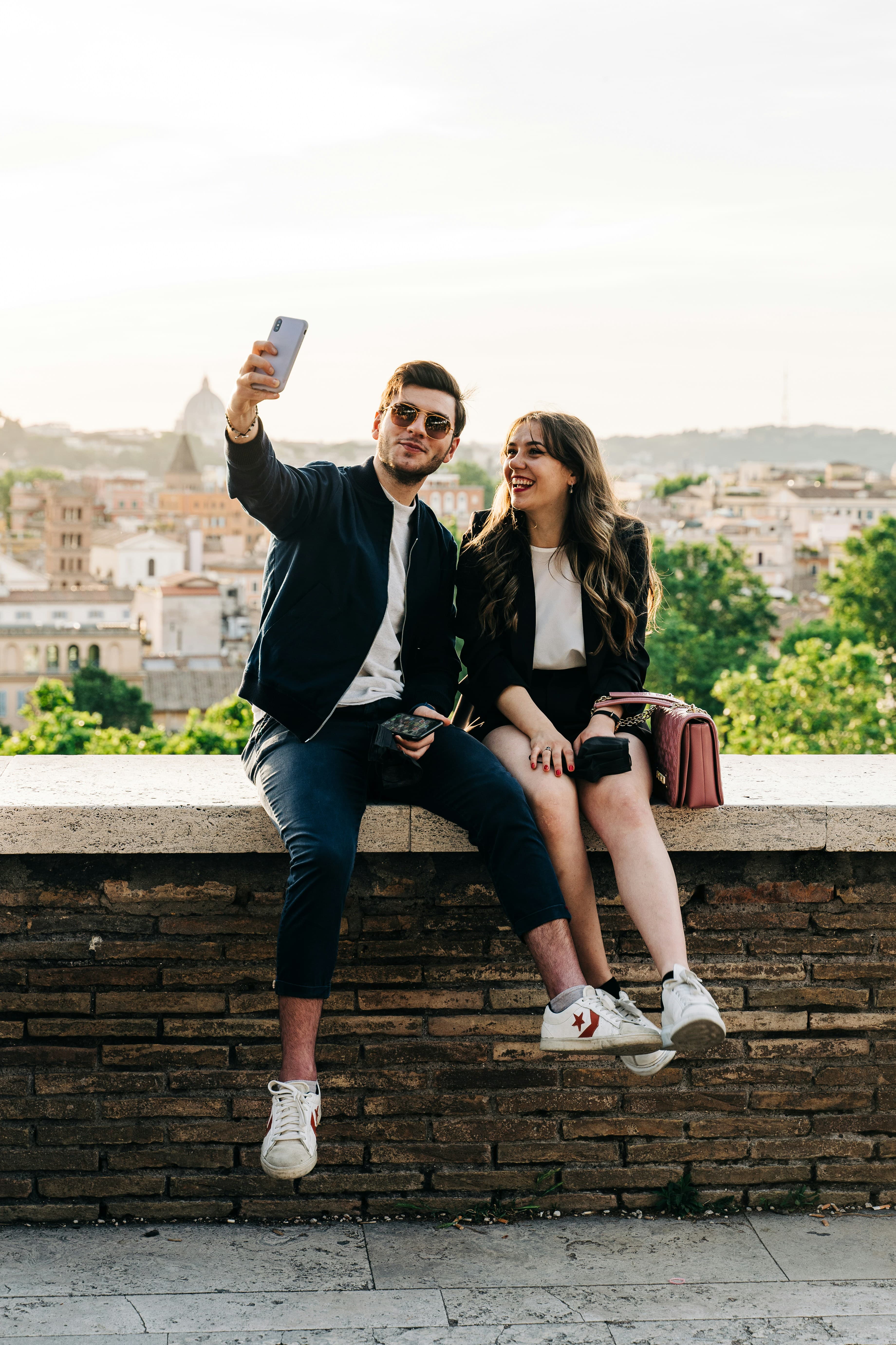 A photo of a young white man and woman sitting on a wall and taking a selfie. Behind them is a view of a European city.