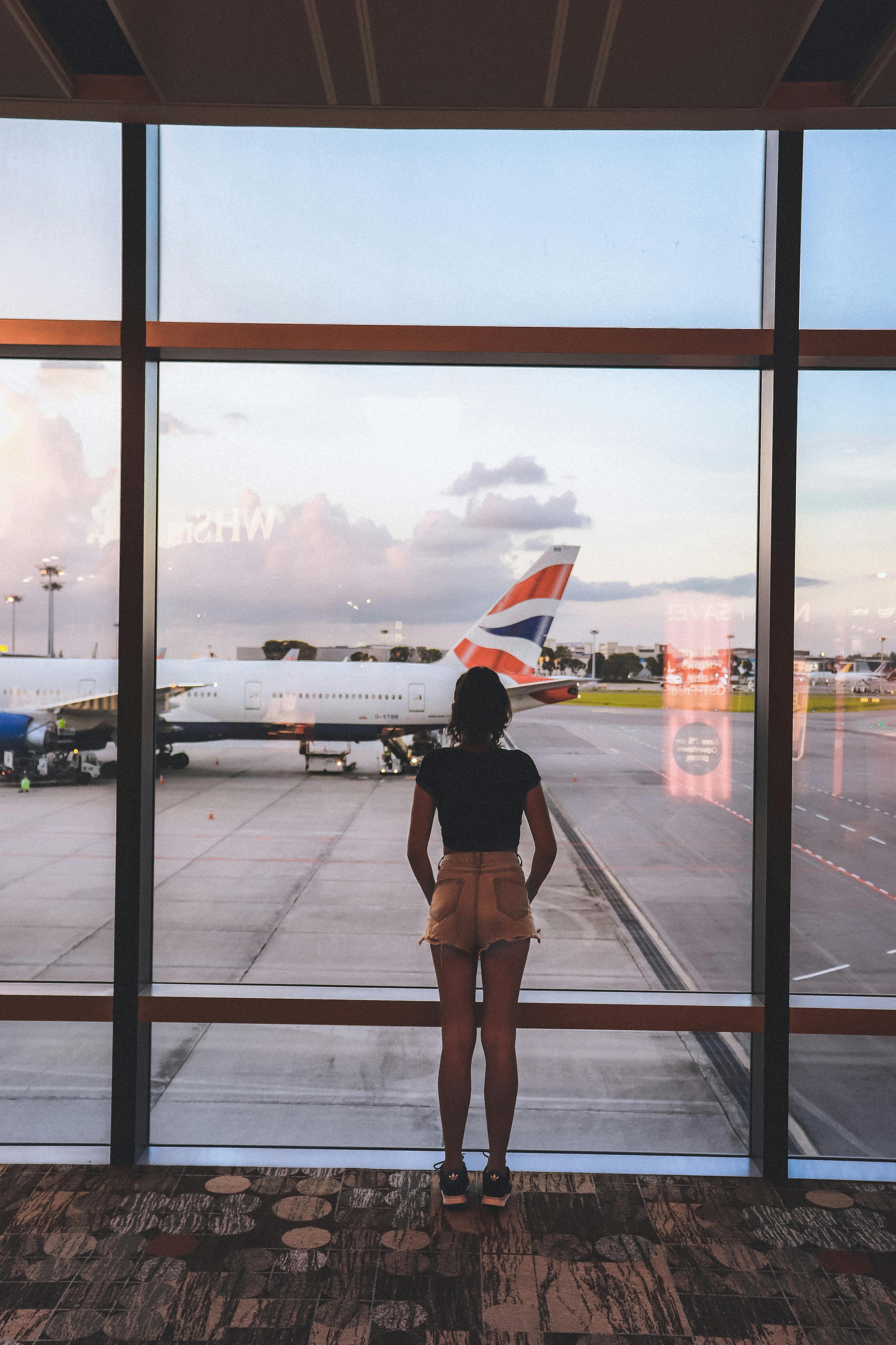 A photo of a young woman taken from behind. She is wearing jean shorts and a dark t-shirt and is looking at an airplane through the window of an airport at dawn.