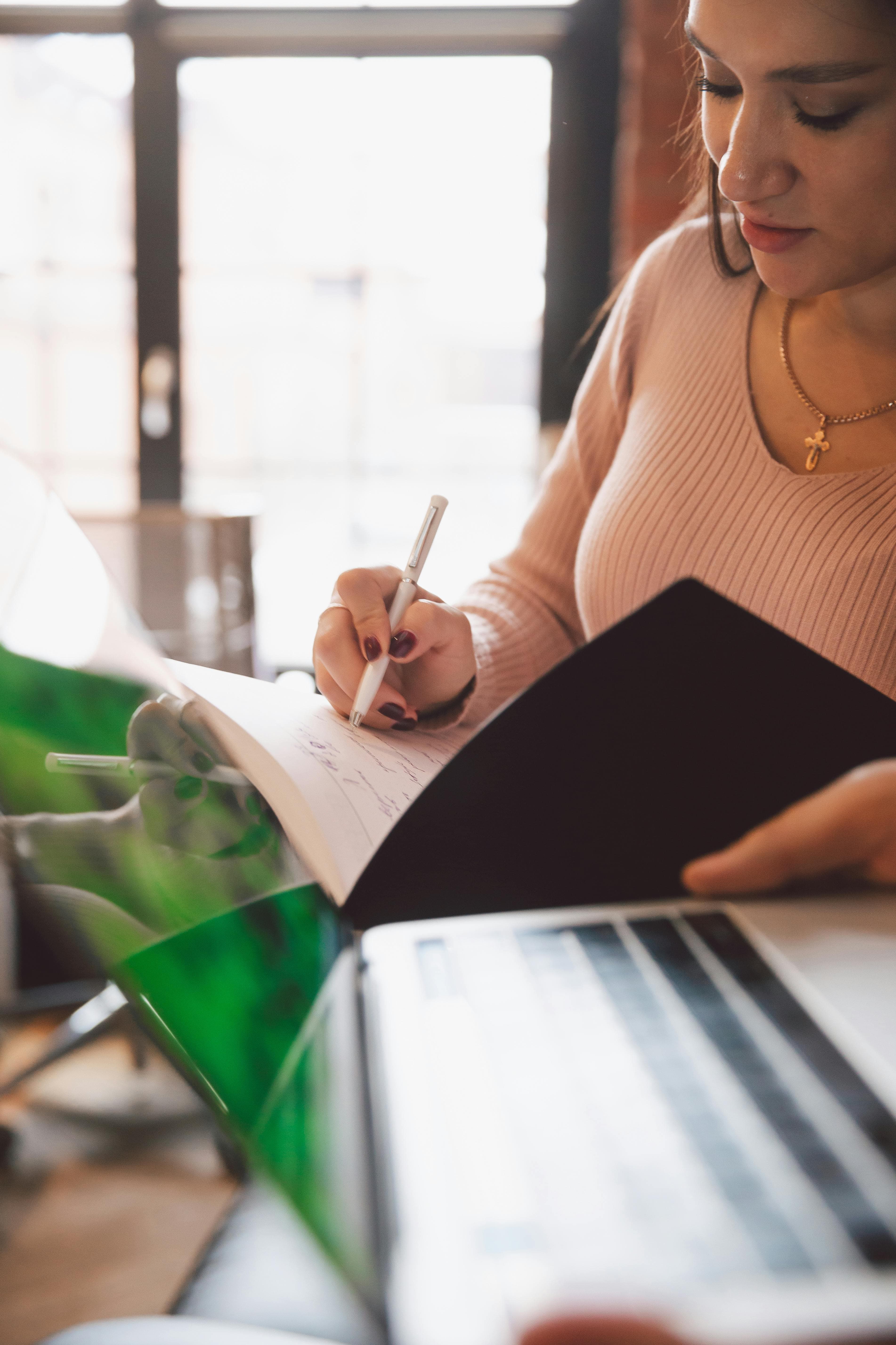 A woman in a pink sweater is writing in a planner or notebook.