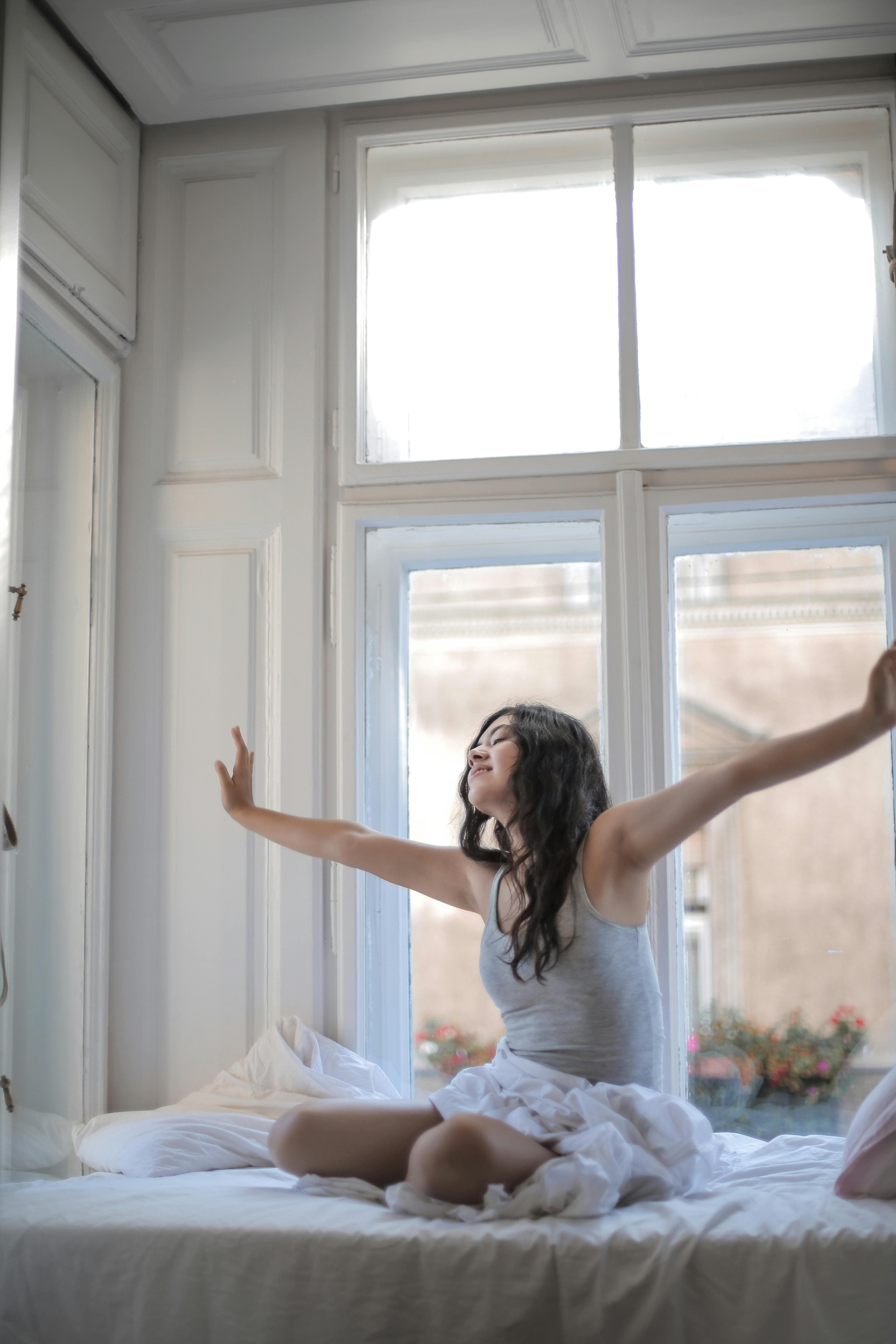 A photo of a woman waking up and stretching in the morning. She is sitting in bed in front of a large urban window.