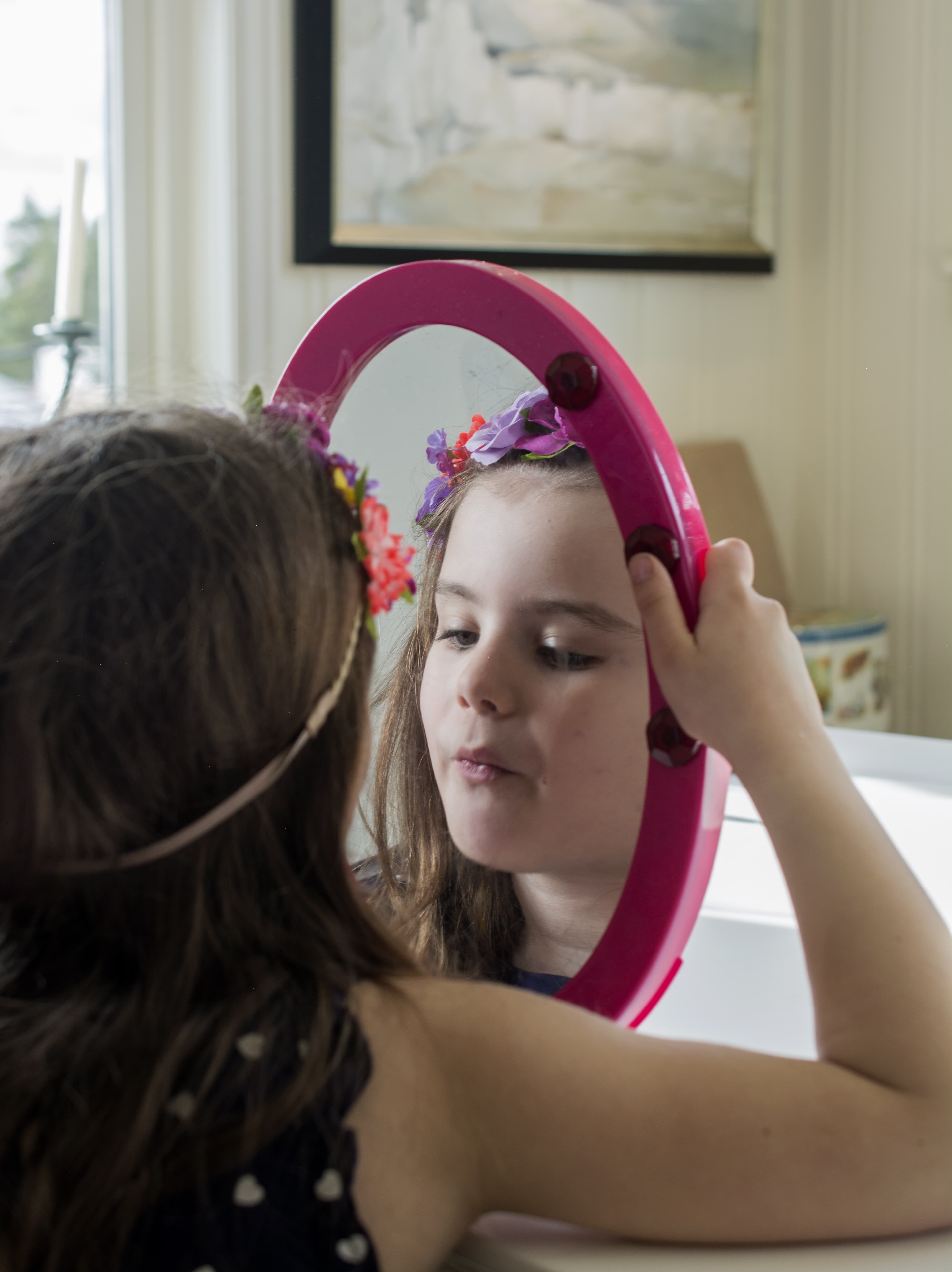 A white girl wearing a flower crown looks at her reflection in a pink-framed oval mirror that she is holding up to her face.