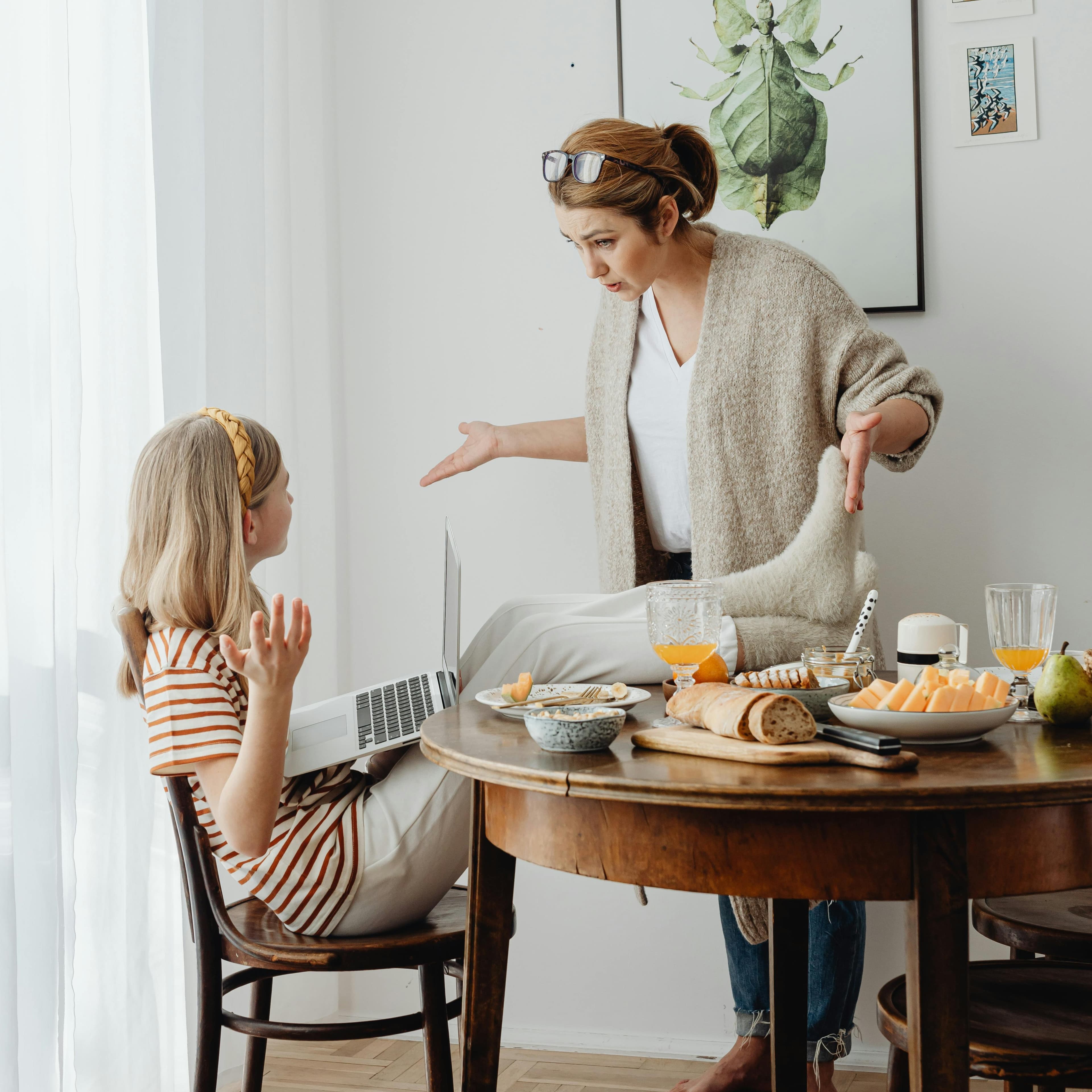 A photo of a pre-teen girl and her mother by a kitchen table with breakfast food. The girl is sitting with her feet on the table and her laptop on her lap. She appears to be arguing with her mother, who is standing behind the table and looks annoyed.
