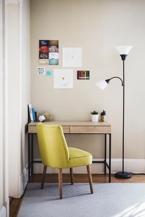 A photo of a desk in a corner. A chair is sitting in front of it. To the right of the desk is a standing lamp. A rug is on the floor. Several pictures are hanging on the wall over the desk. On the desk are several items, left to right: some books, a decorative paperweight, a plant, and a cup of pens.