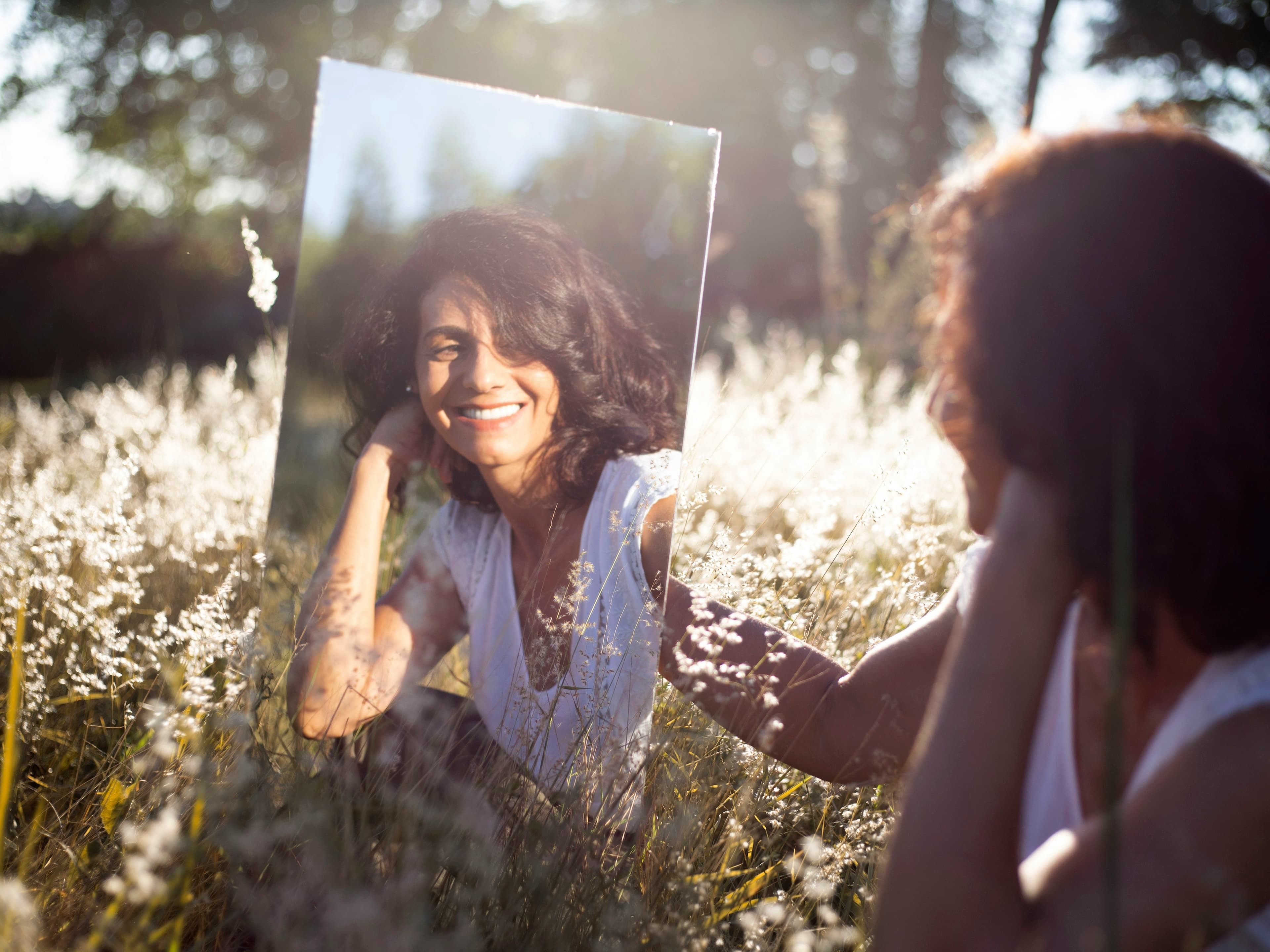 A photo of a woman with brown skin, dark curly hair, and a white t-shirt. She holds up a mirror to look at her reflection in a sunny field of grass.