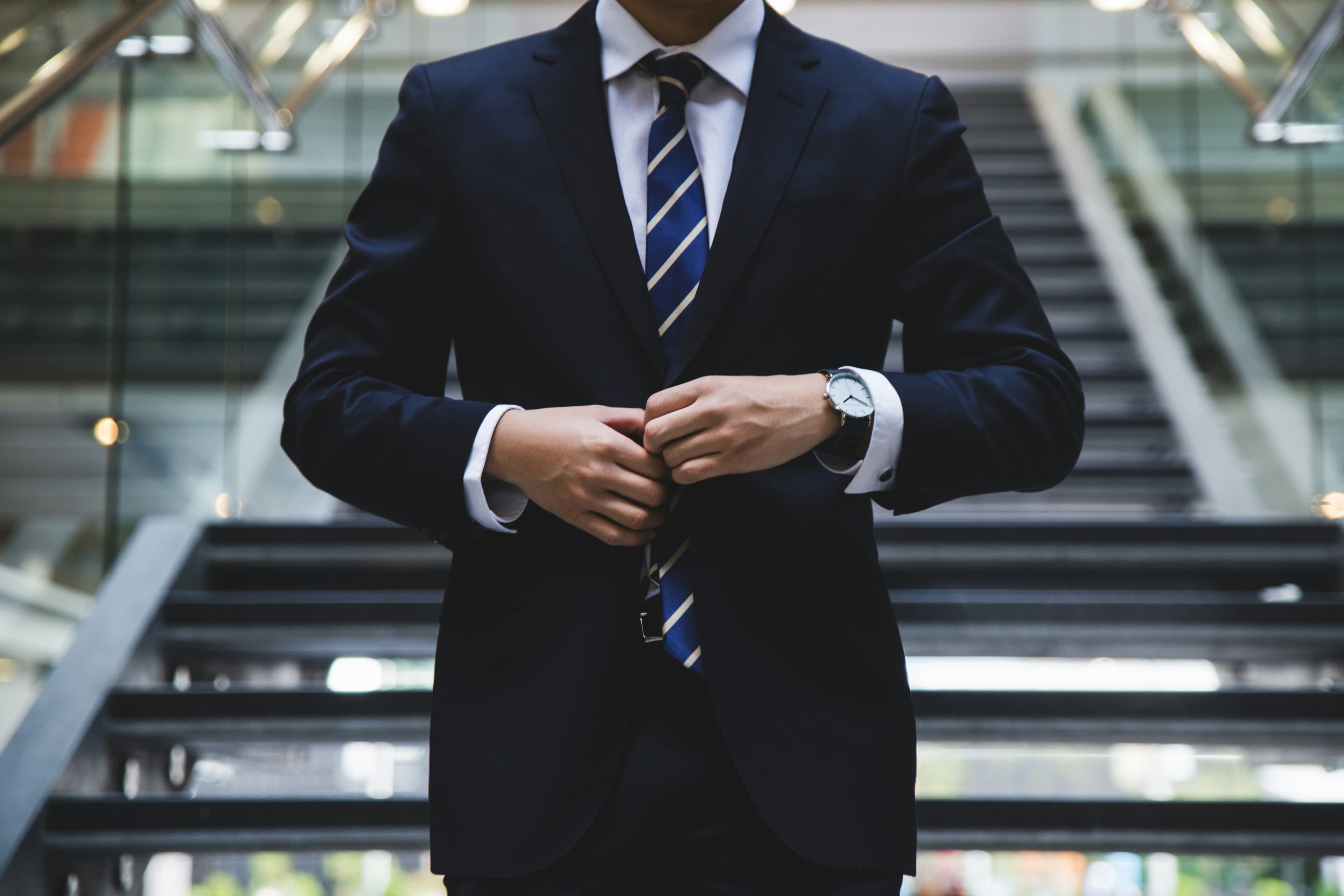 A photo of the torso of a white man in a navy blue suit and tie, with a modern staircase in the background.