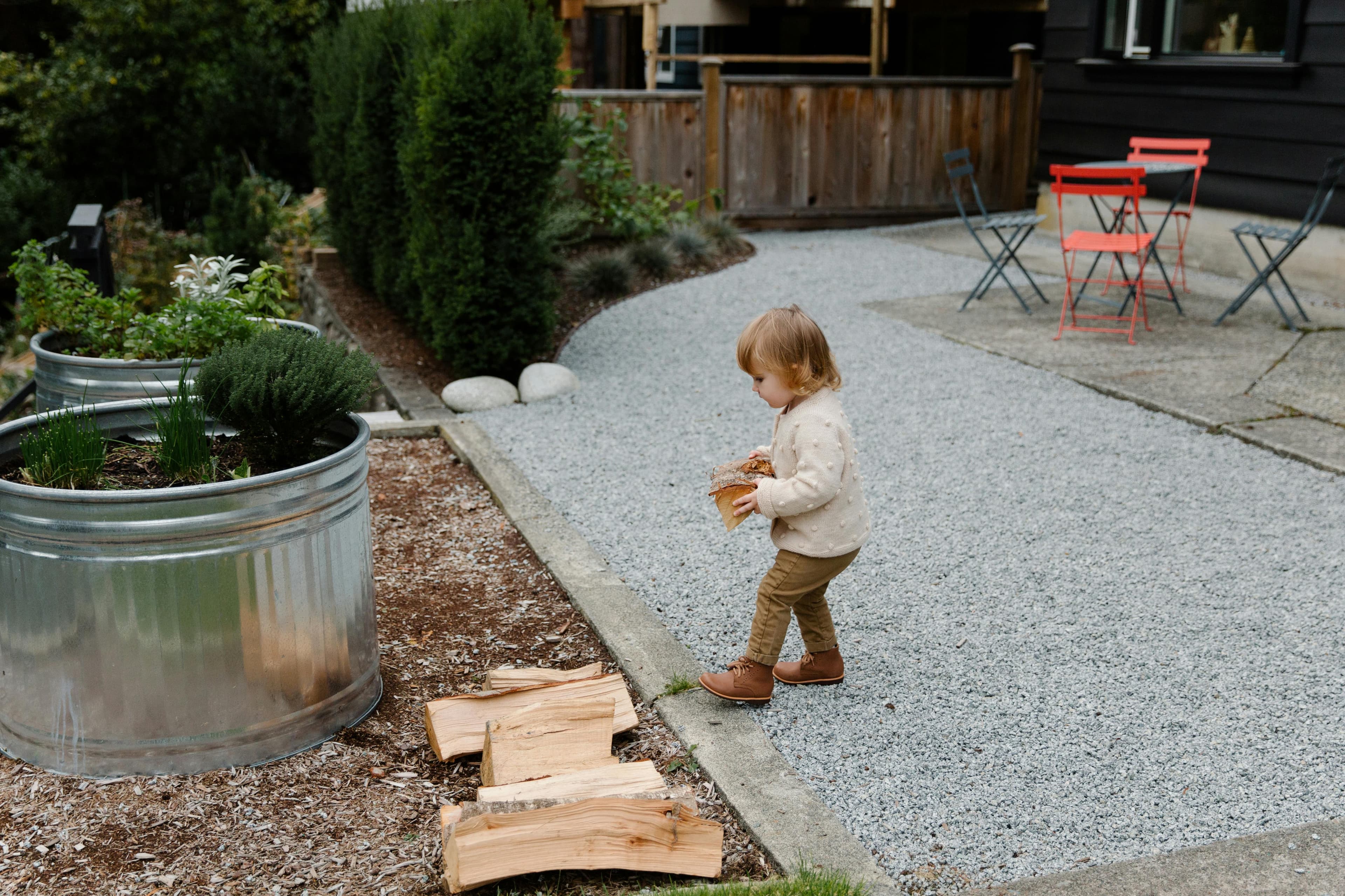 A photo of a little blonde kid working to carry pieces of firewood from one end of the patio to the other. She has carried several pieces already, and is about to set down another one on the pile.