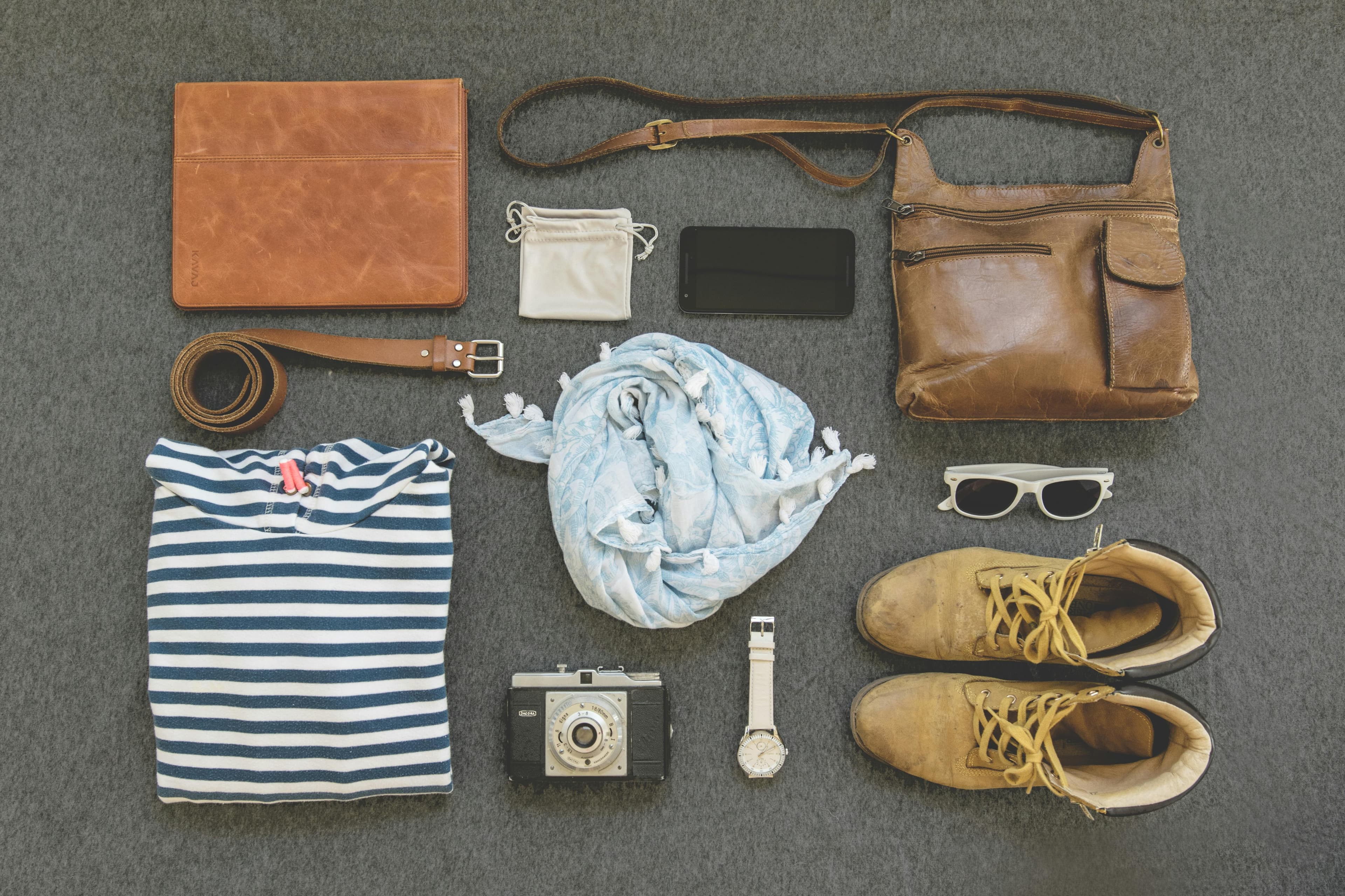 A photo of several items on the floor. A pair of boots, a pair of sunglasses, a purse, a cell phone, a coin purse, a scarf, a watch, a camera, a folded shirt, a belt, and a tablet in a leather case