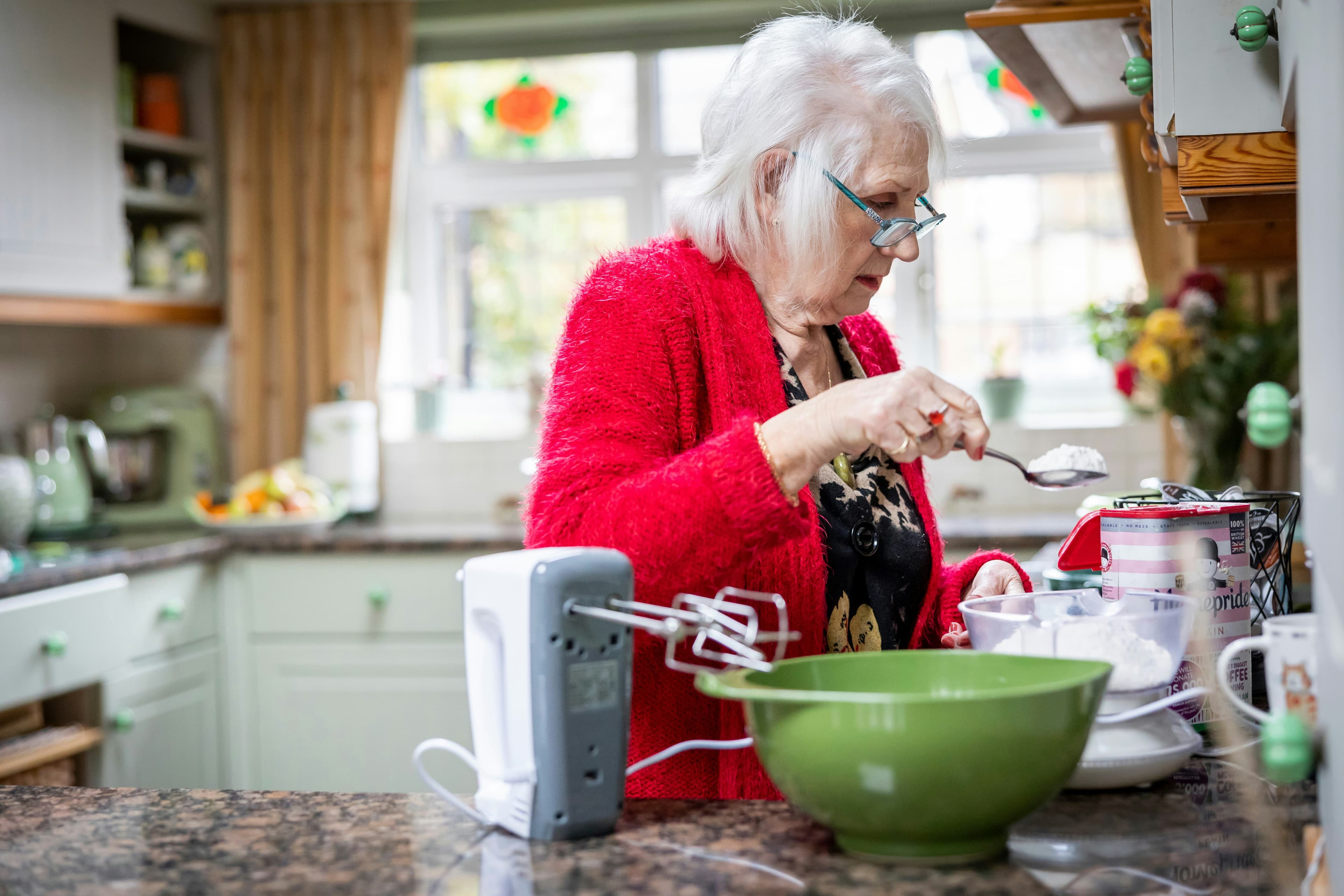A photograph of an elderly white woman with white hair and a red sweater baking in a kitchen. She is measuring flour into a green mixing bowl using a spoon.