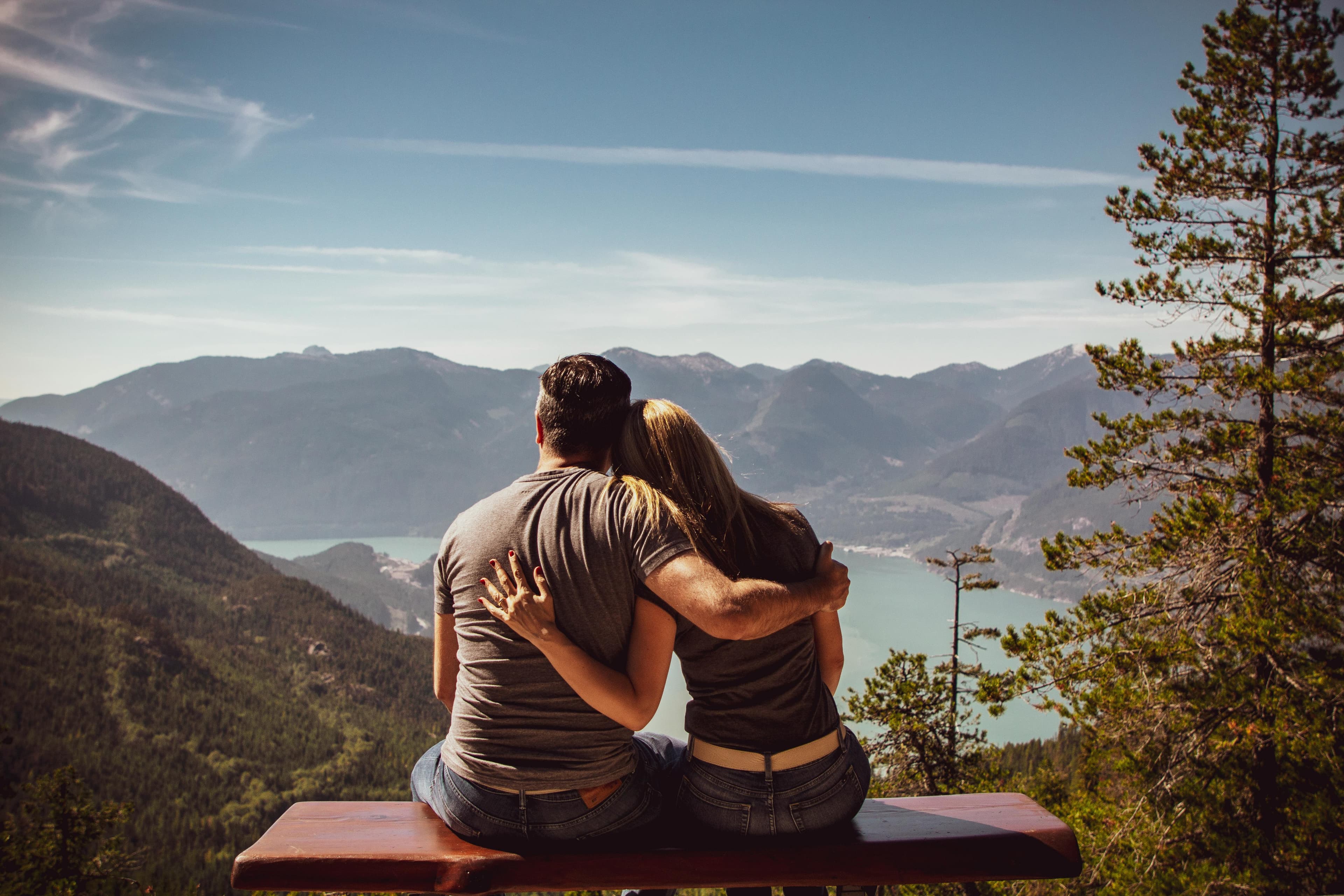 A photo taken from behind a man and a woman with their arms around each other. They are sitting on a picnic table, looking out at a mountain view.