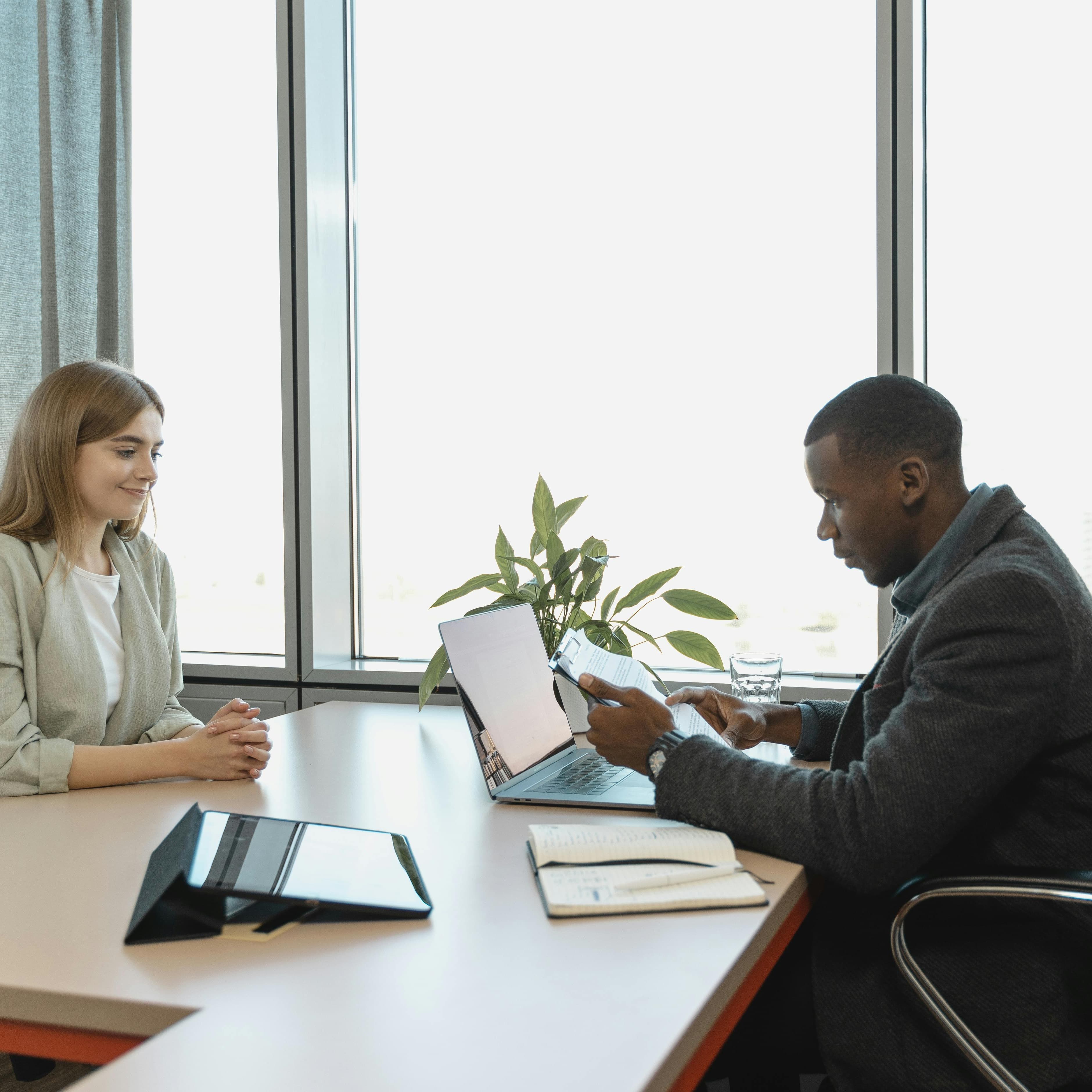 A job interview. A man in an office is sitting at a desk, looking over some paperwork while a woman on the other side of the desk is smiling.