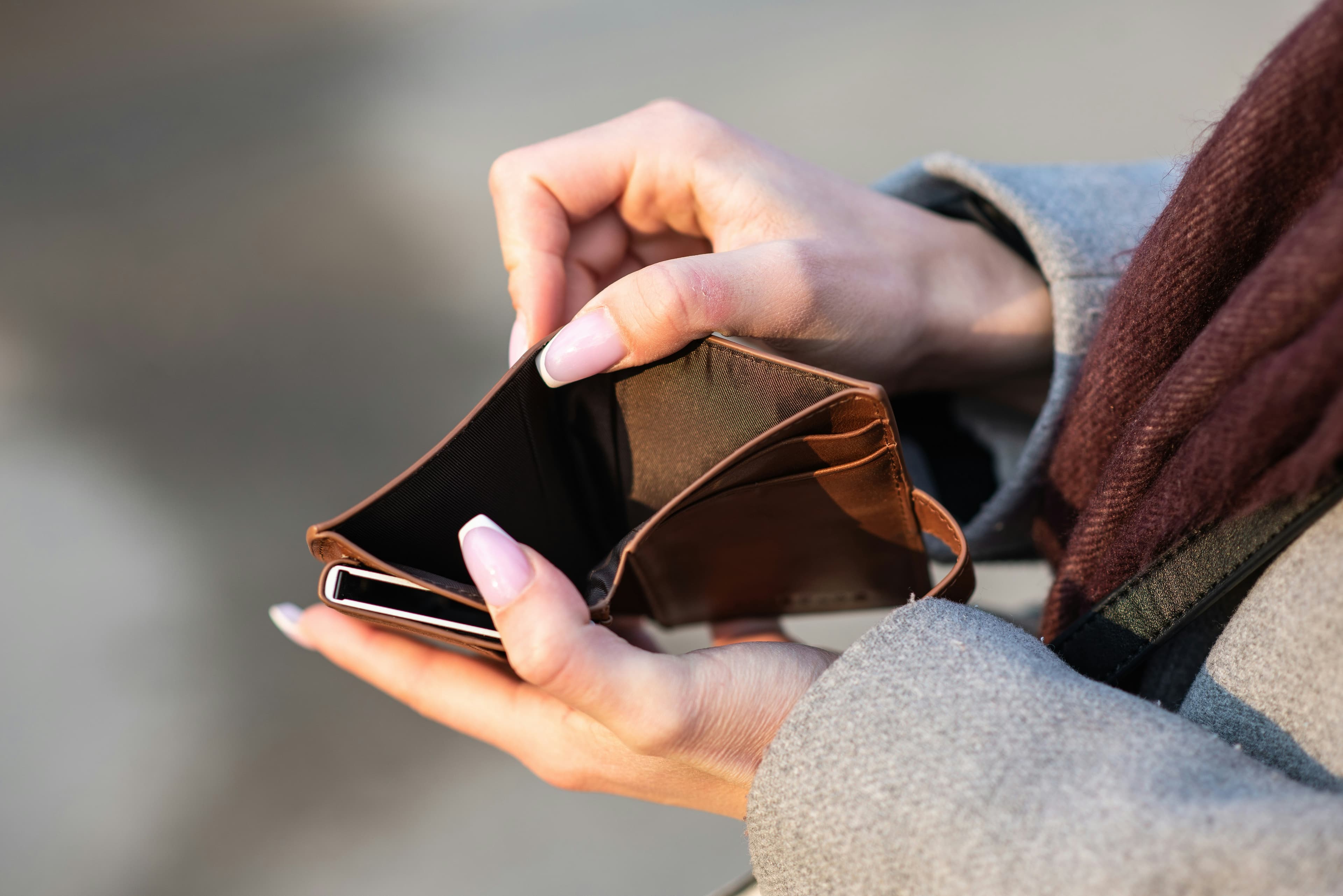 A photo shows the hands of a white woman in a wool winter coat and scarf as she holds open an empty brown wallet.