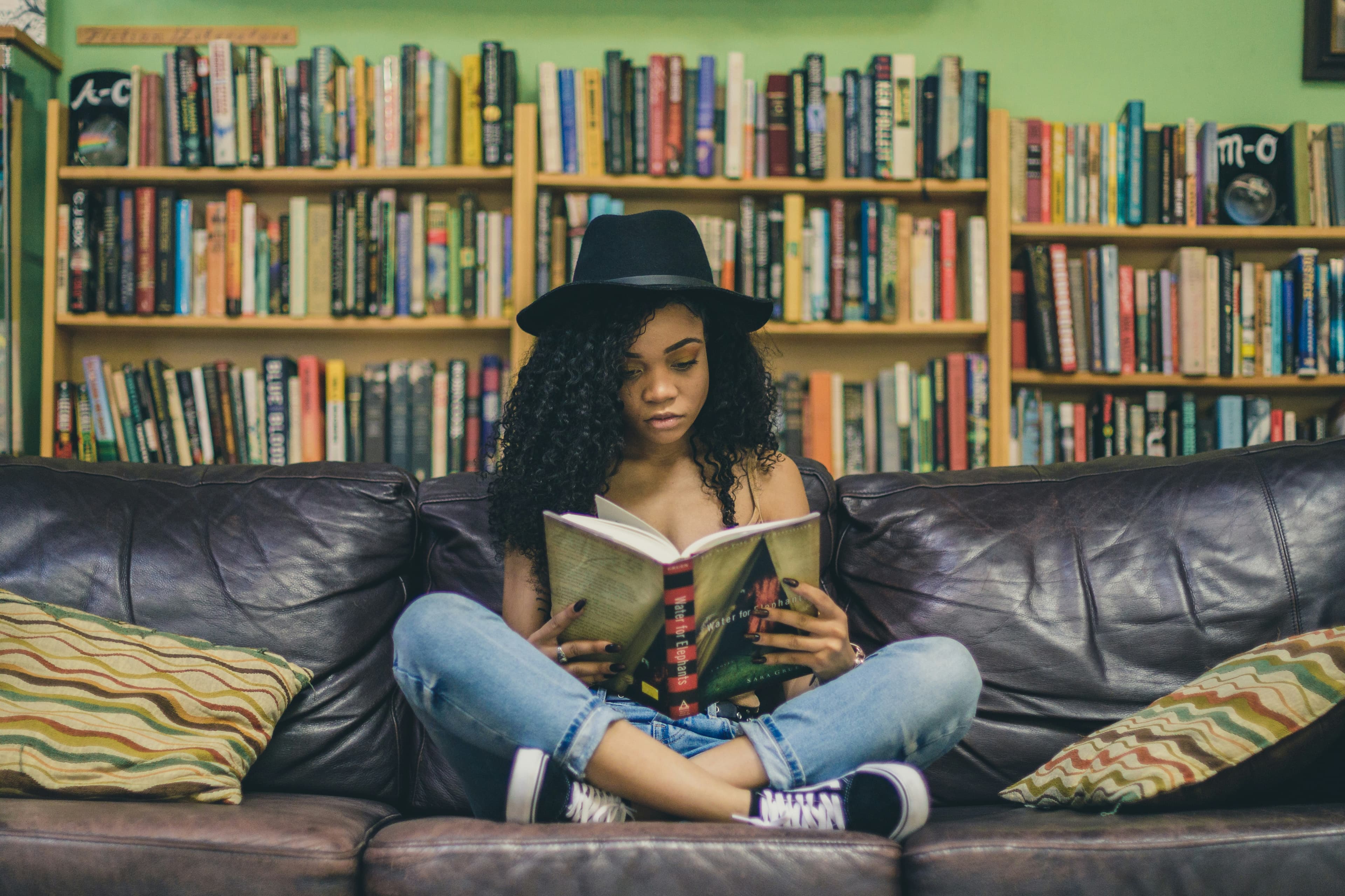 A photo of a mixed-race woman in a fedora sitting cross-legged on a black leather couch reading a copy of "Water for Elephants." Behind her are several shelves packed with books.