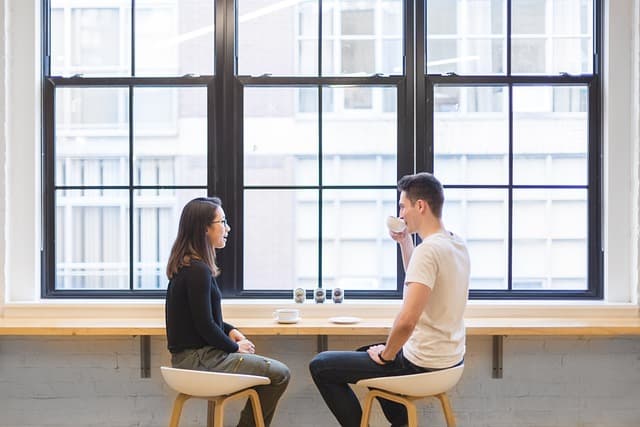 A young man and woman sit at a breakfast bar in front of a second-story window in a modern building.