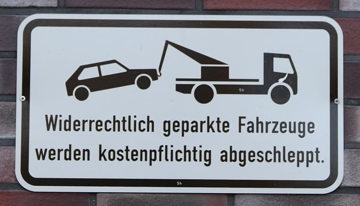 An image of a street sign with a car being towed. The German text reads: "Widerrechtlich geparkte Fahrzeuge werden kostenpflichtig abgeschleppt."