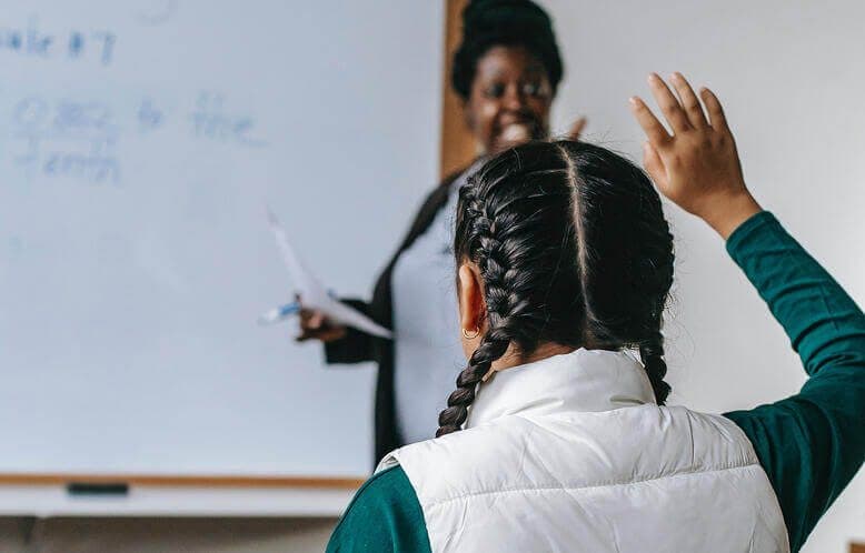 A young girl in classroom with her hand risen to ask a question
