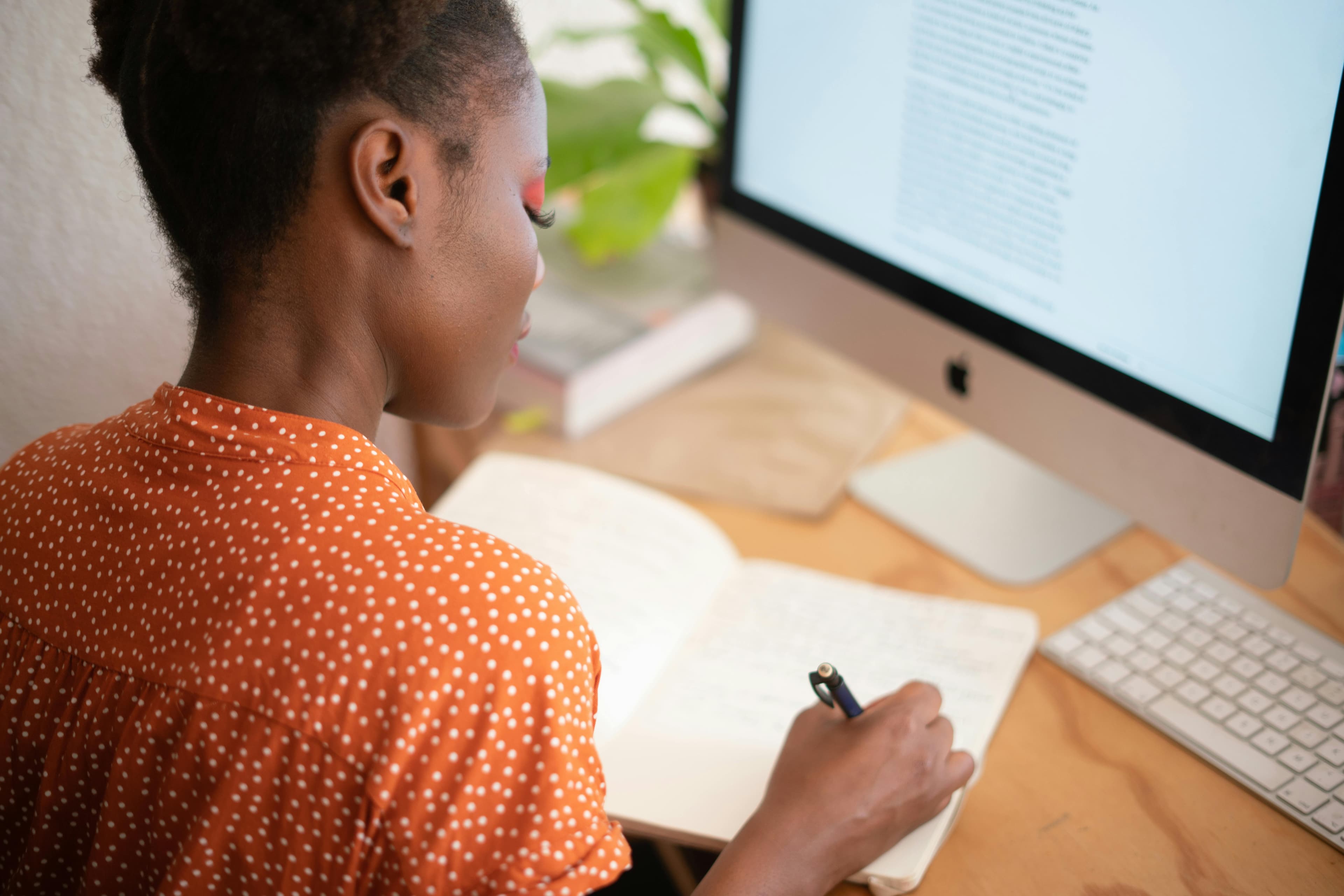 A photo of a woman sitting in front of a computer, writing with a pen in a notebook.