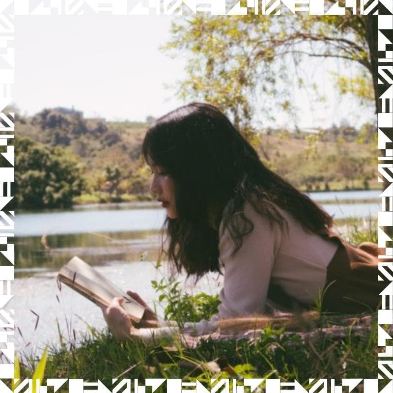 A girl reading a book by a body of water next to a tree