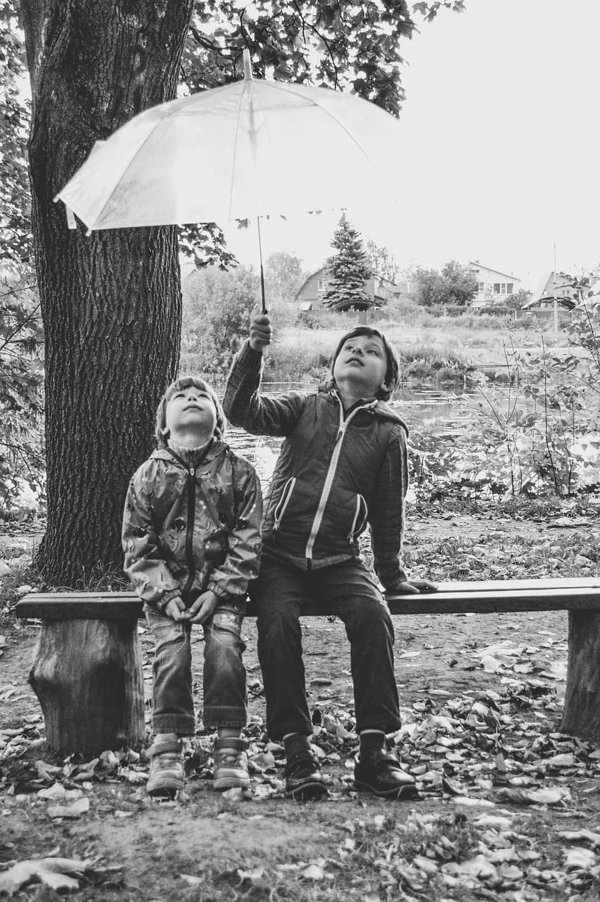 An old black-and-white photo of two boys in raincoats sitting on a bench holding an umbrella.