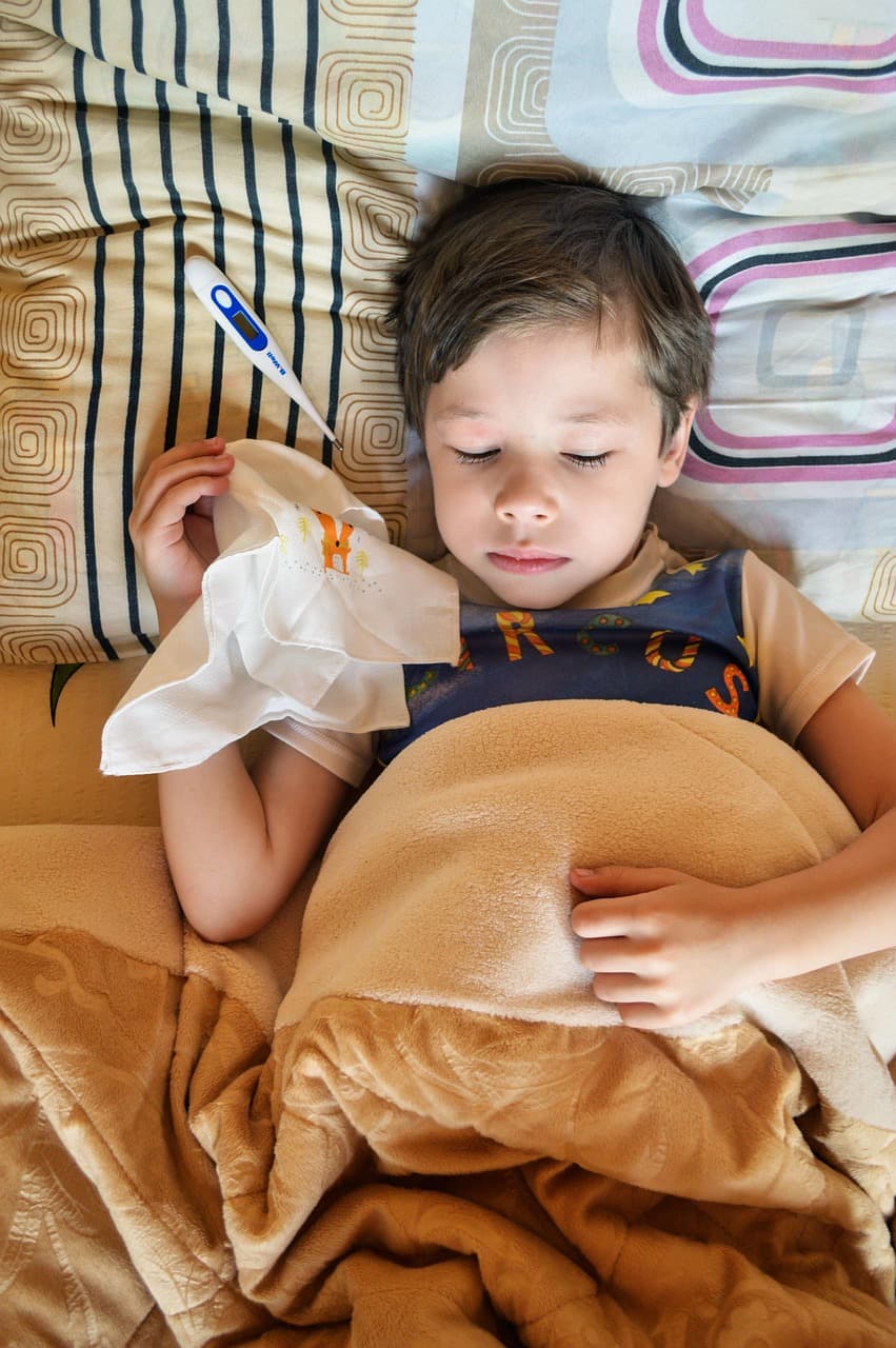 A kid lying in bed, sick, with a thermometer by his head and a handkerchief in his hand.