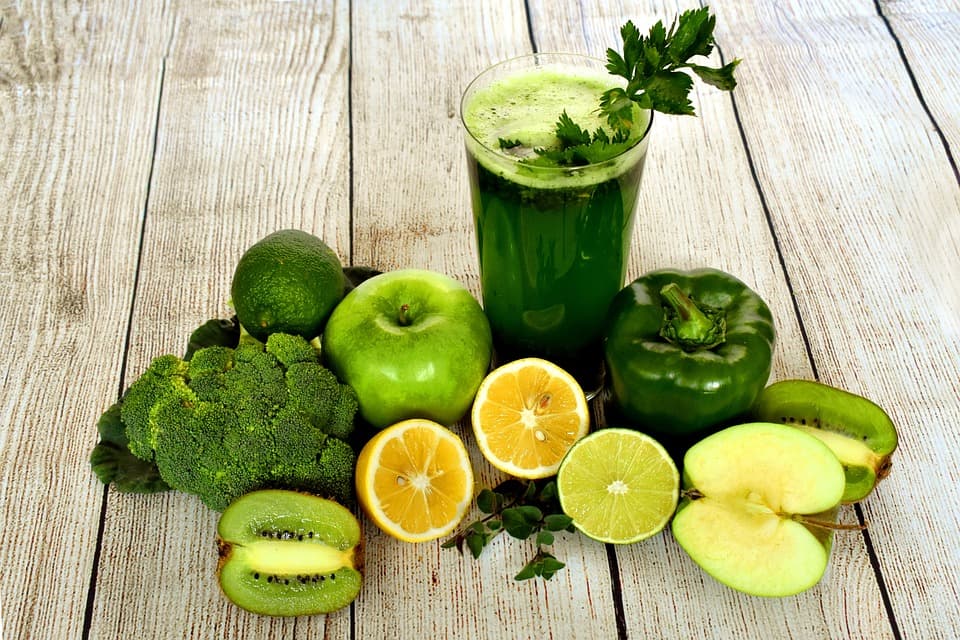 A photo of a bunch of bunch of green fruits and veggies. 1 bunch of broccoli, 1 glass of green juice, 2 halves of a kiwi, 1 whole lime, 1 whole apple, 1 half an apple, 1 green pepper, 2 halves of a lemon, 1 half of a lime.