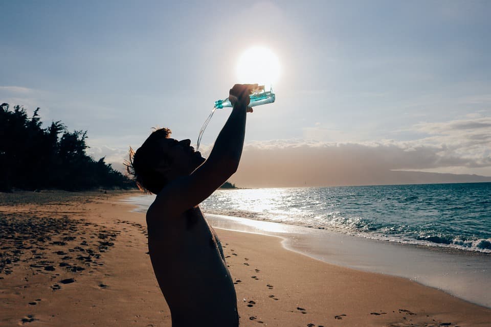 A photo of a man on a beach. He is silhouetted against the sun as he raises a water bottle up in the air and pours the water into his mouth.