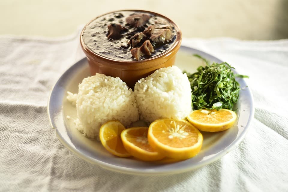 A plate of food on a white linen tablecloth. The plate contains a small bowl of brown stew, two balls of rice, some greens, and a sliced citrus fruit.