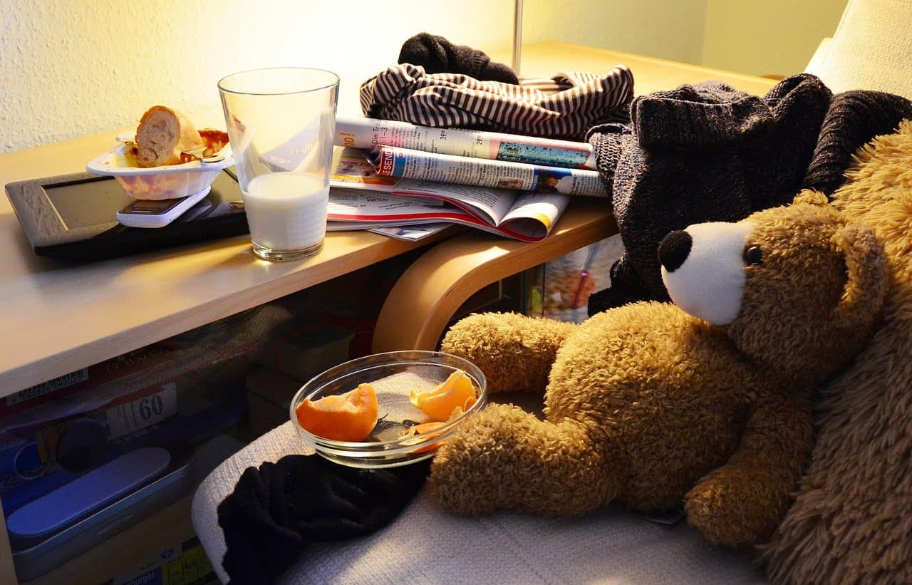 A photo of a teddy bear sitting on a couch. In front of the teddy bear, on the couch, is a bowl of fruit sitting on top of a sock. On the side table, a bowl of bread is sitting on top of a cell phone, which is on top of a tablet. On the table, there is also a stack of a magazines with some clothes on top of them. In front of everything on the table is a glass of milk.