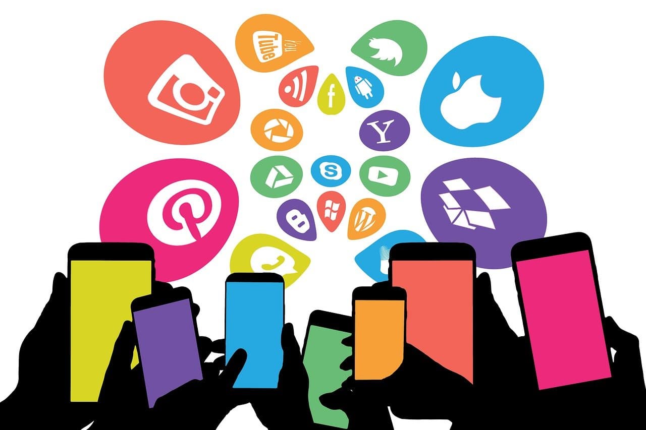 A clipart graphic of several hands holding up smart phones that glow different bright colors. Above them the logos of various social media and other web-based companies float in multicolored bubbles.