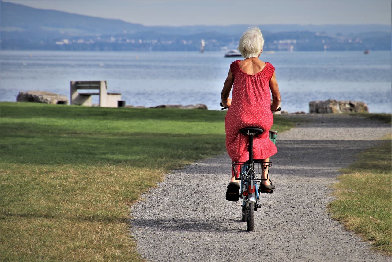 A photo of an older lady in a dress riding a bike through a park by the water.