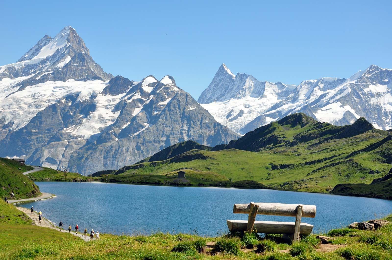In Switzerland, tall snow-capped mountains stand behind green hills. Hikers walk along a blue lake. There is a rough-hewn wooden bench in the foreground.
