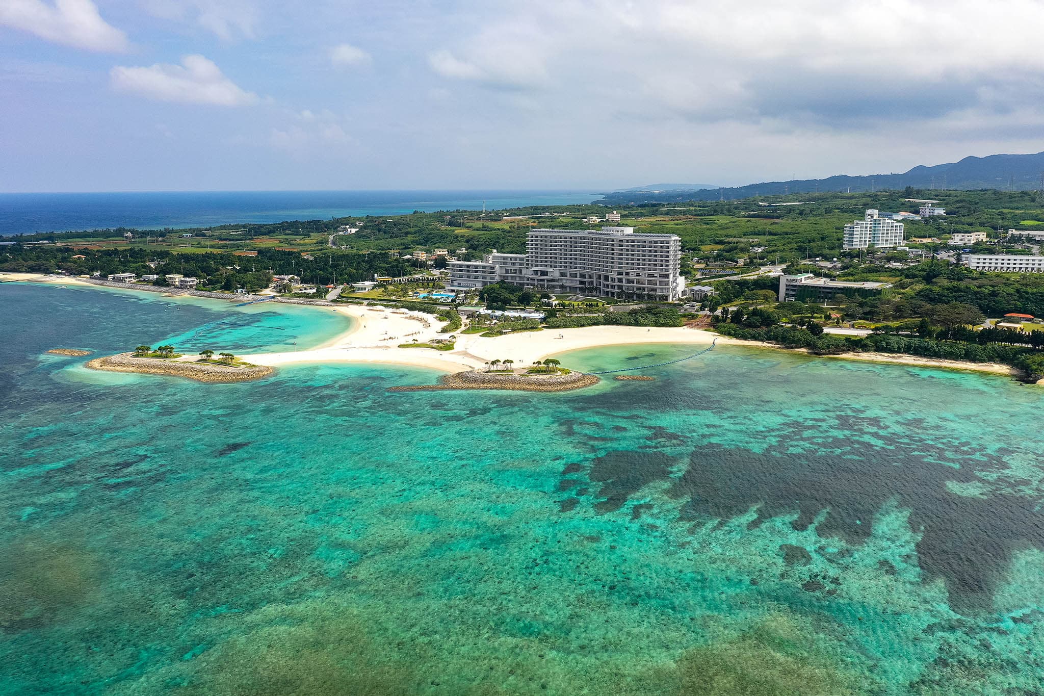 An aerial shot of the clear turquoise waters off the coast of Okinawa. There are some large buildings, but most of the landscape is green and healthy.