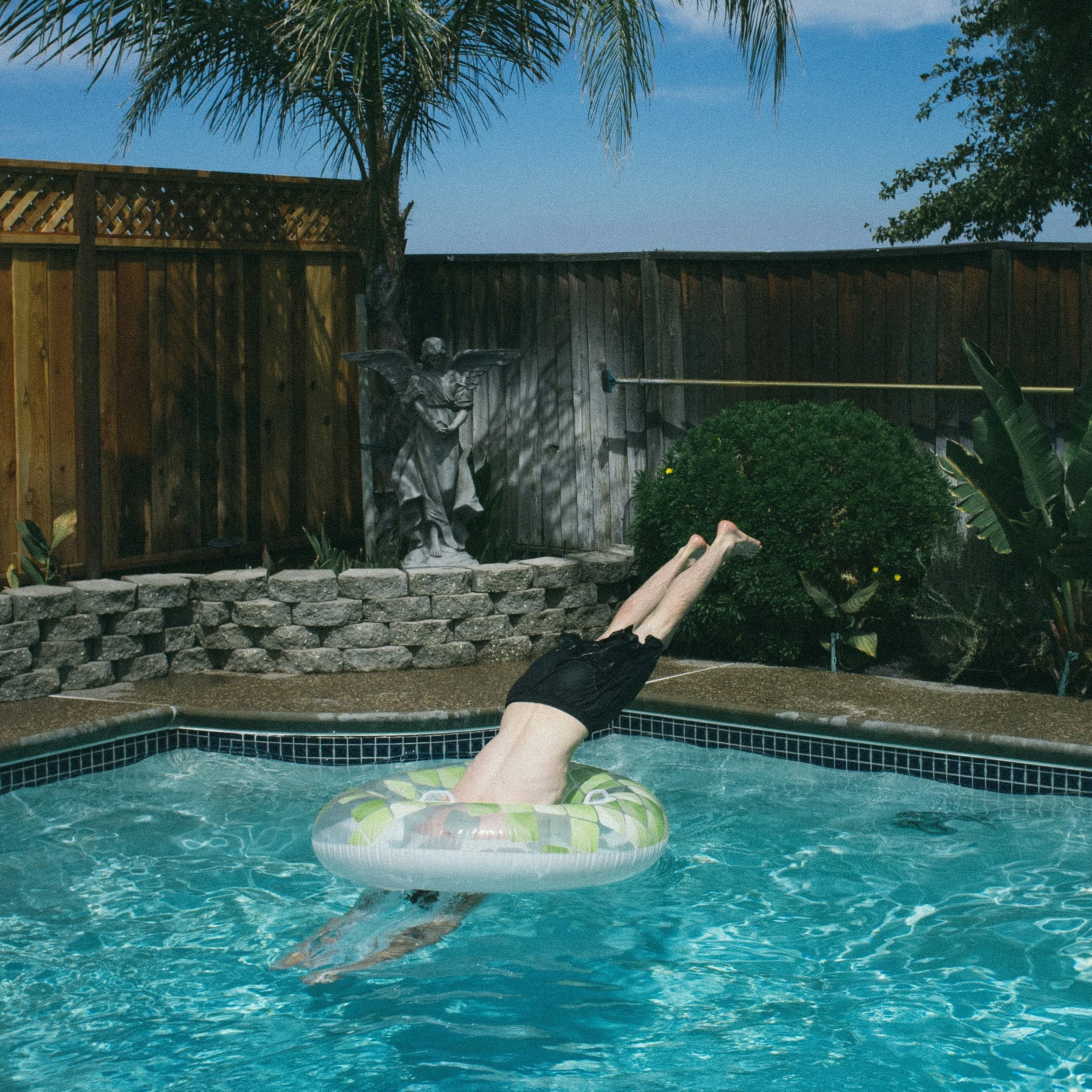 A photo of a man diving into a swimming pool, through an inner tube floating on the water. The pool appears to be in a backyard, as it's surrounded by privacy fencing, and it is surrounded by a narrow garden.