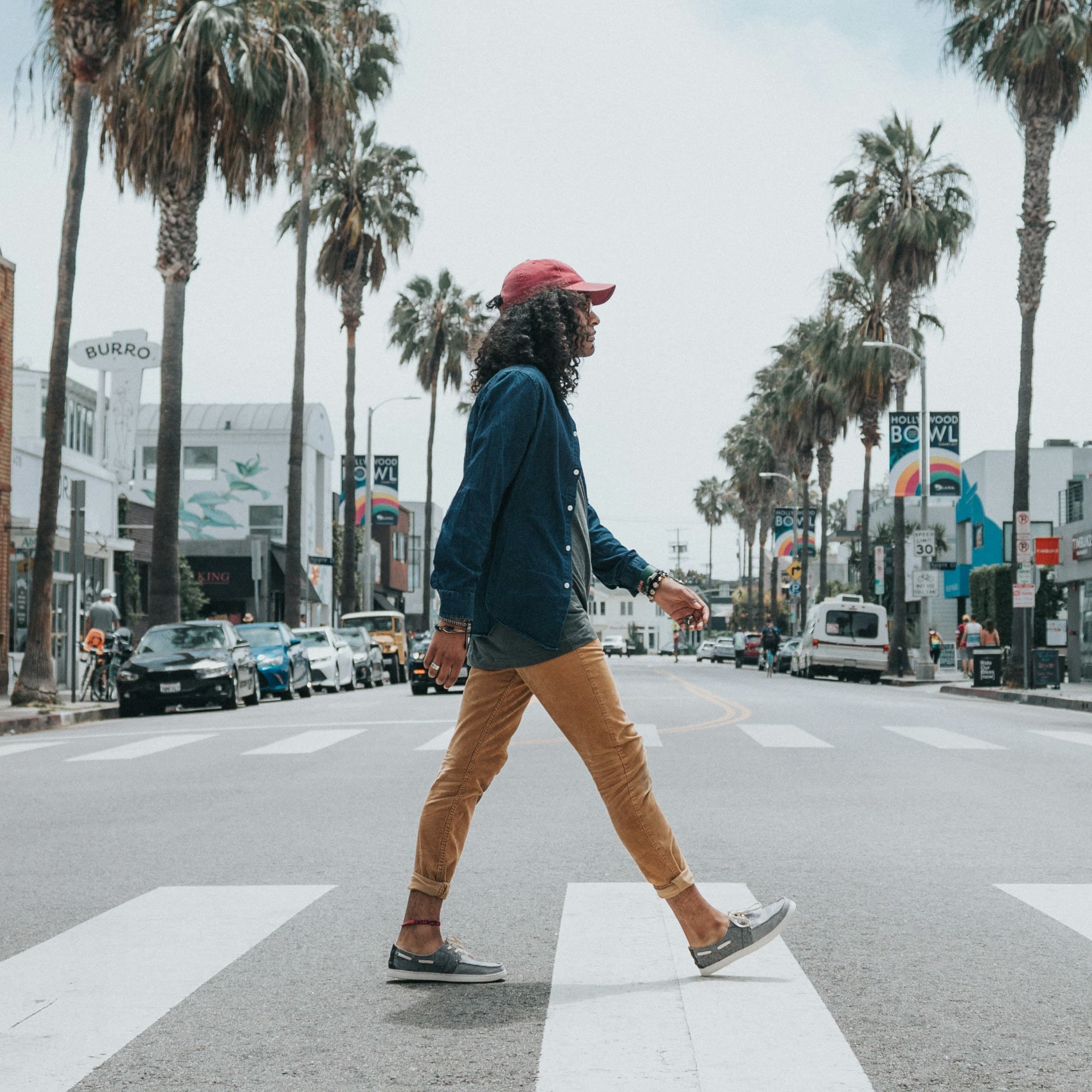 A man with shoulder-length curly hair, a blue dress shirt, gray t-shirt, tan chinos, boat shoes, and a red baseball cap is crossing the street at a zebra crosswalk. He is currently in the middle of the road. Behind him we can see that the street is lined with palm trees and looks to be an older downtown from a Western city, perhaps in California or the Southwest.