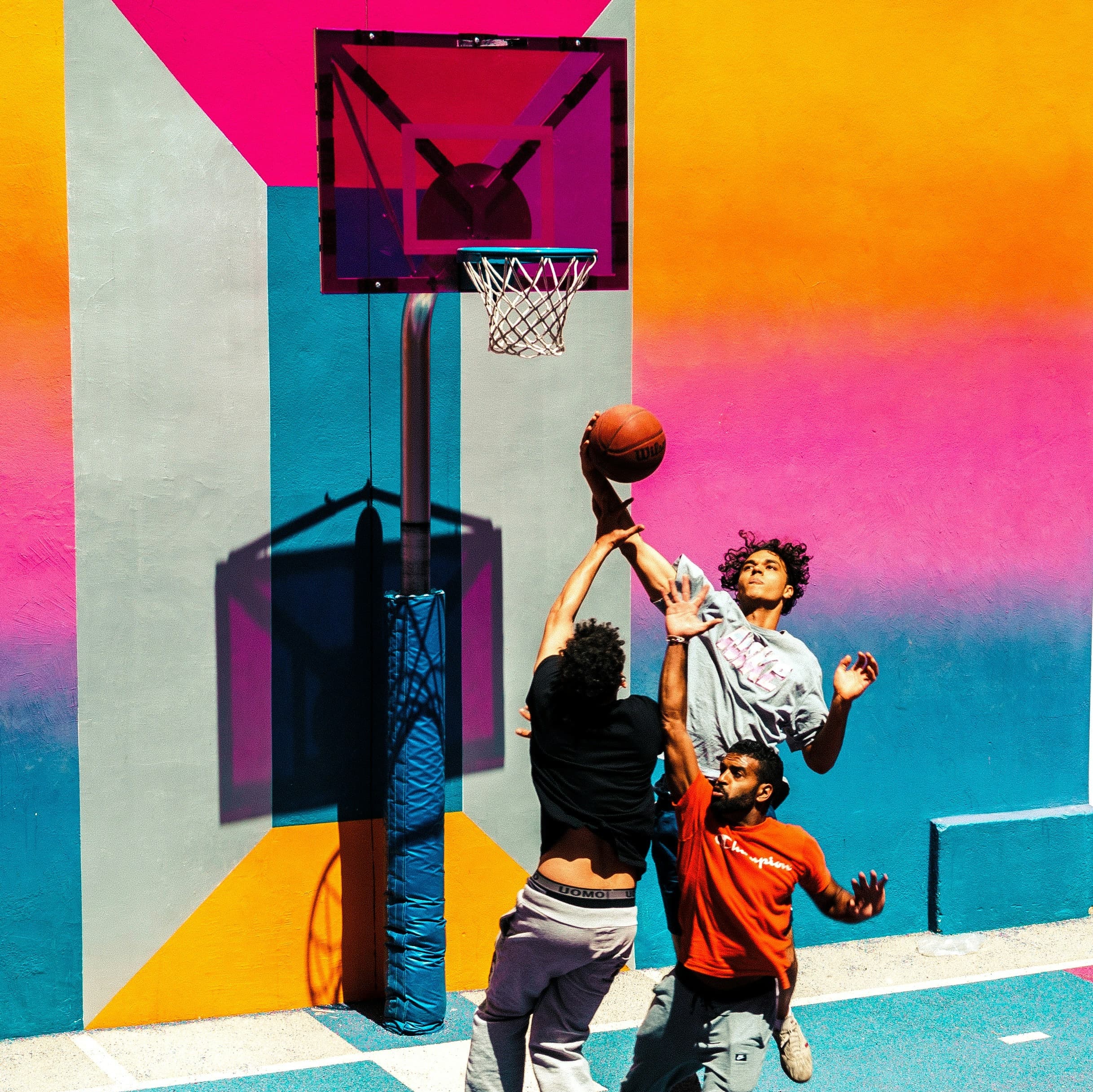 Three young men of different races are playing basketball in front of a concrete wall painted with a colorful design. The photo has caught them all jumping into the air. One young man is going to shoot a basket (or perhaps dunk the ball) while the other two are reaching out to try to interfere with the shot.