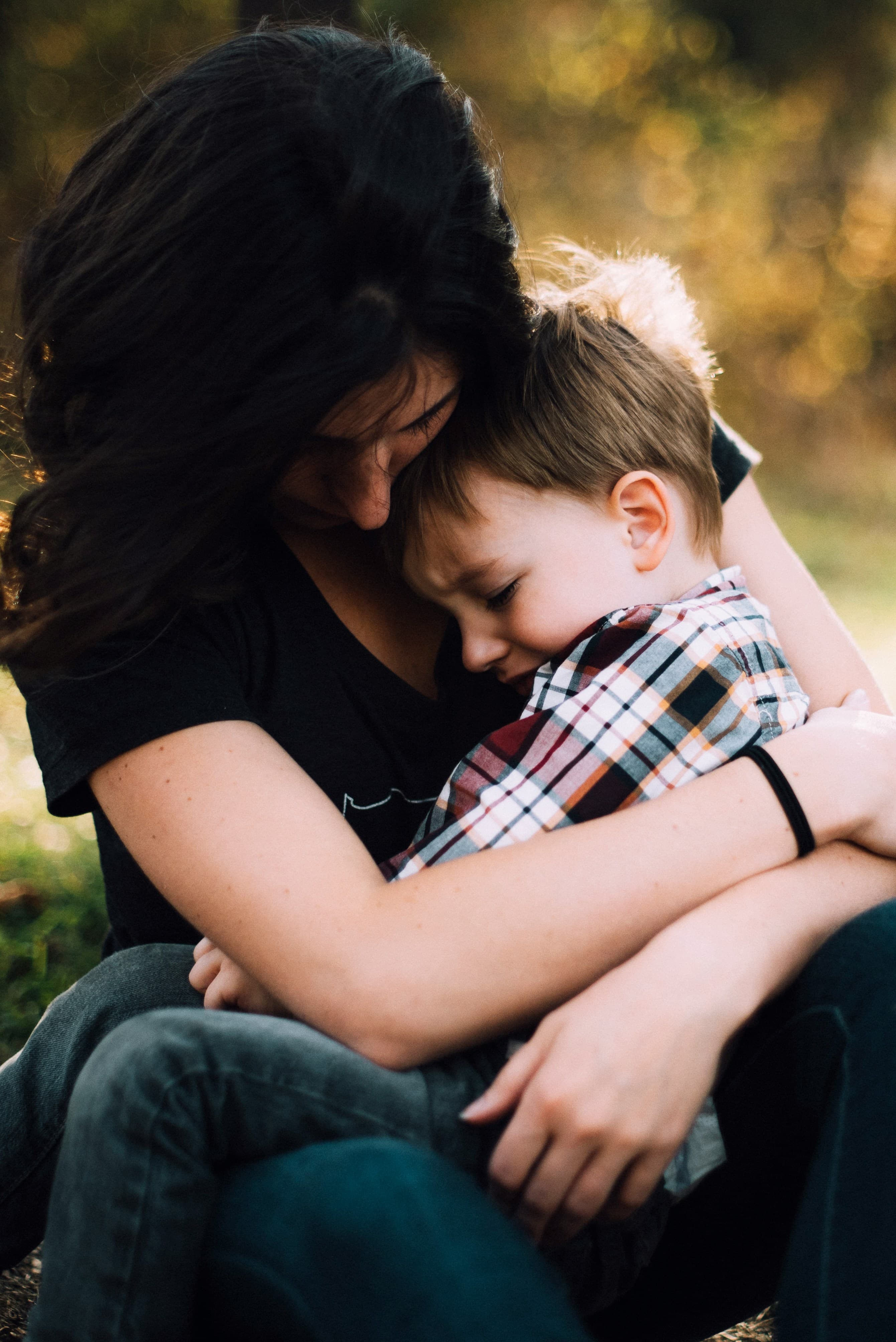 A woman with dark hair holds a young blond boy on her lap and hugs him as he cries.