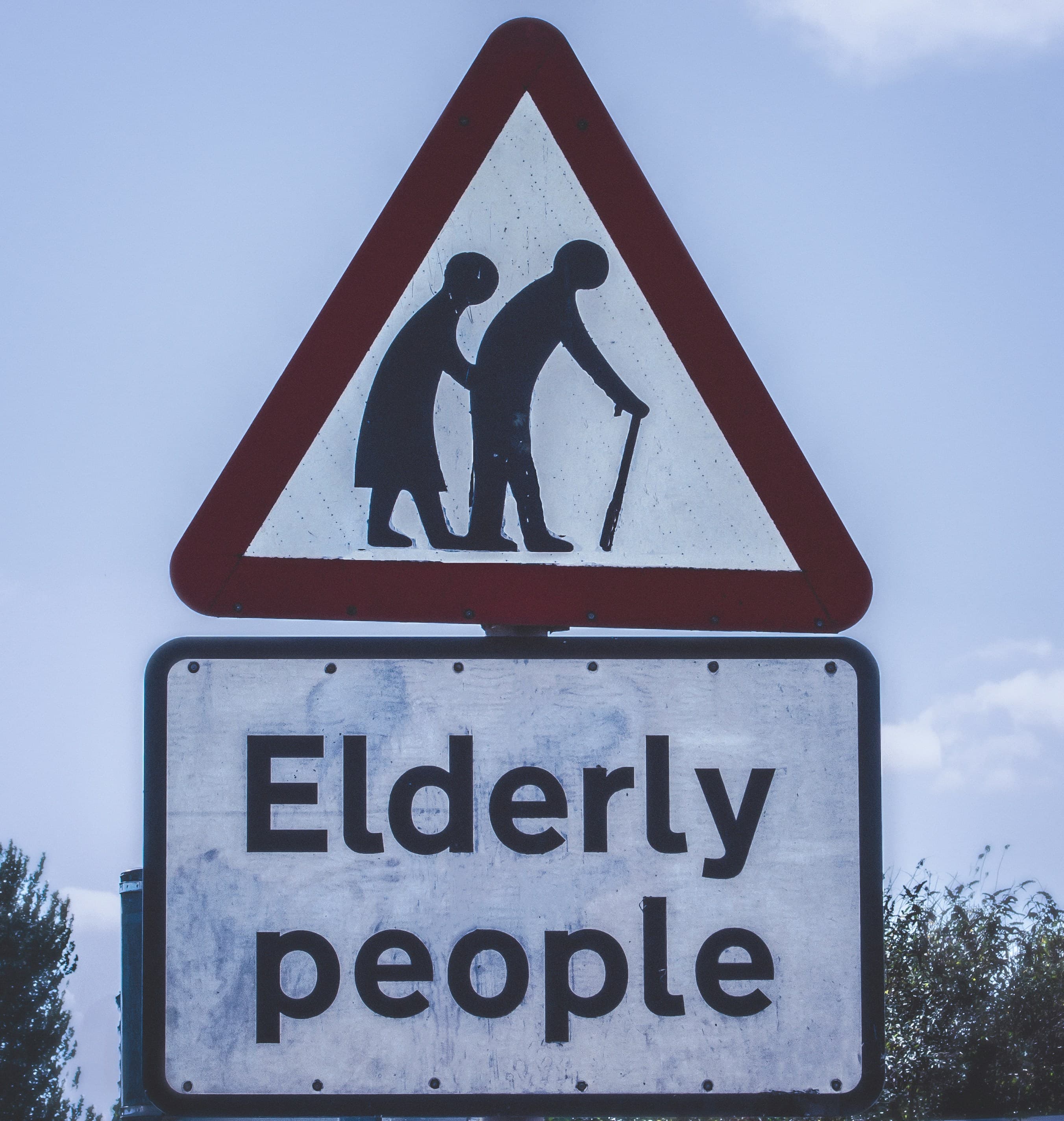 A triangular sign over a rectangular one. The triangular sign has a red border and a graphic of two hunched people, on of whom has a cane. The bottom sign contains text: "Elderly people"