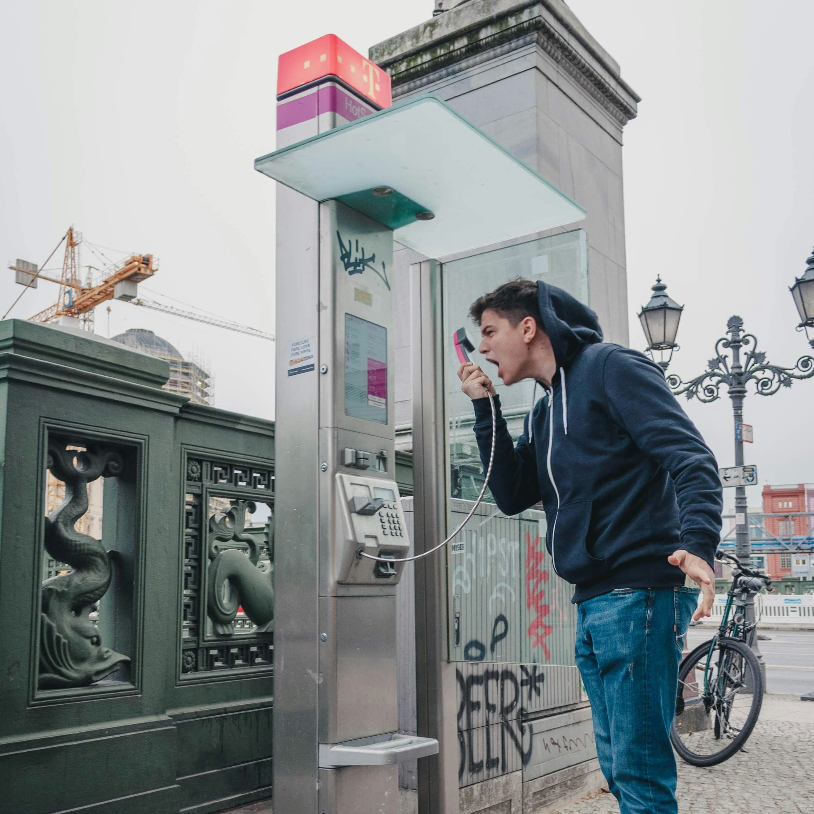 A photo of a white man in a dark hoodie screaming angrily into the receiver of a pay phone. He holds the receiver in front of his mouth instead of up to his ear. The pay phone appears to be in a public square, likely in a European city.