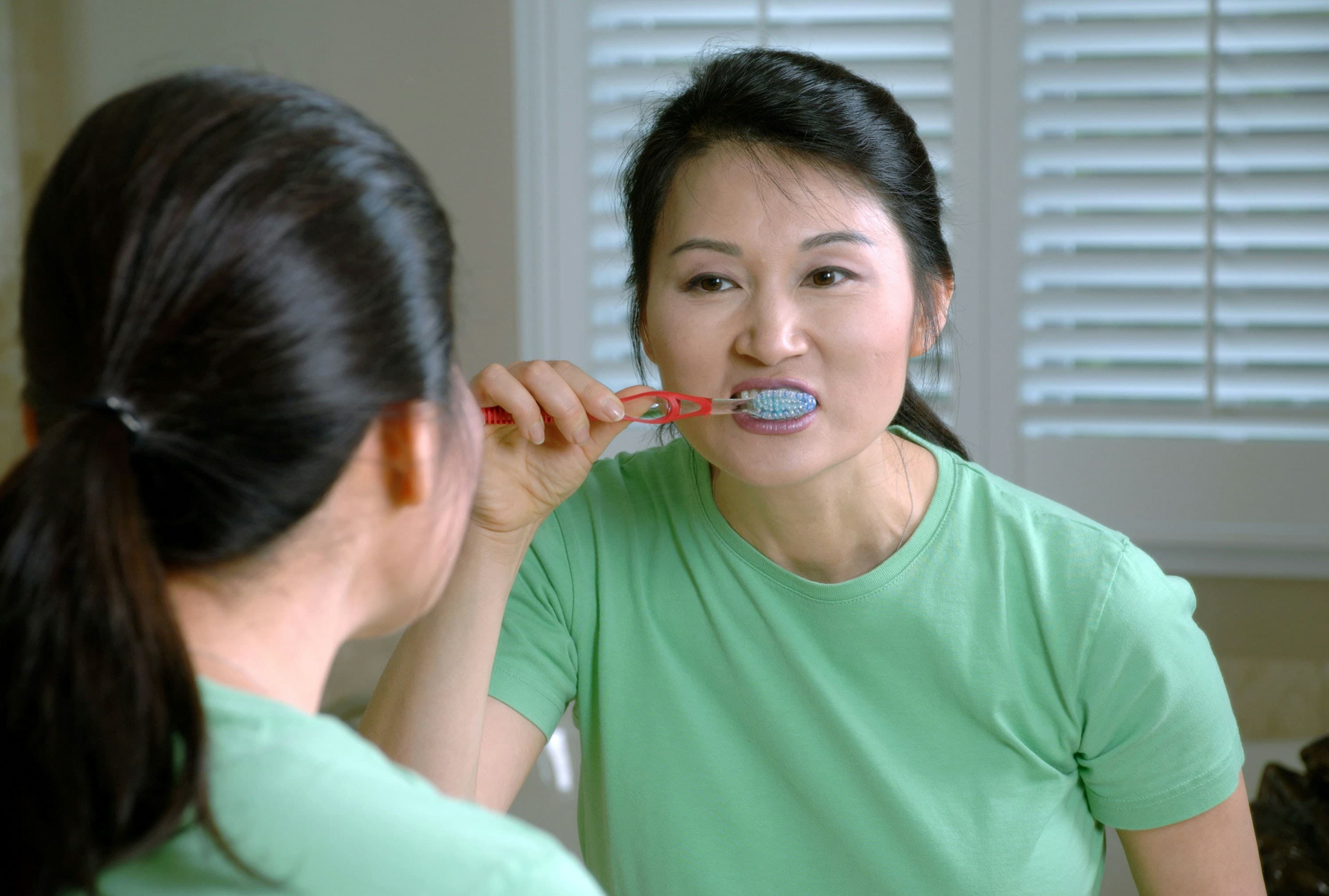 A photo of a woman brushing her teeth, looking at her reflection in the mirror.