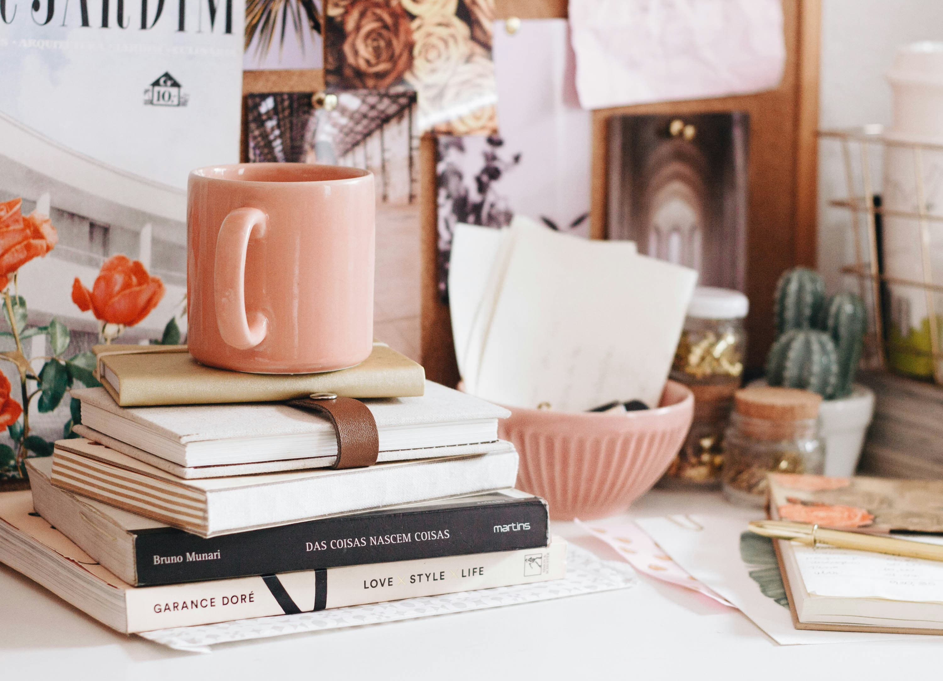 A photo of someone's workspace with a lot of brown, white, and warm pink. We see a pink coffee mug on top of a stack of books, a pink bowl full of letters, a small cactus, several jars, an open notebook, some loose papers, and, in the back, a bulletin board leaned up against the wall with several photos of architecture and flowers pinned to it.