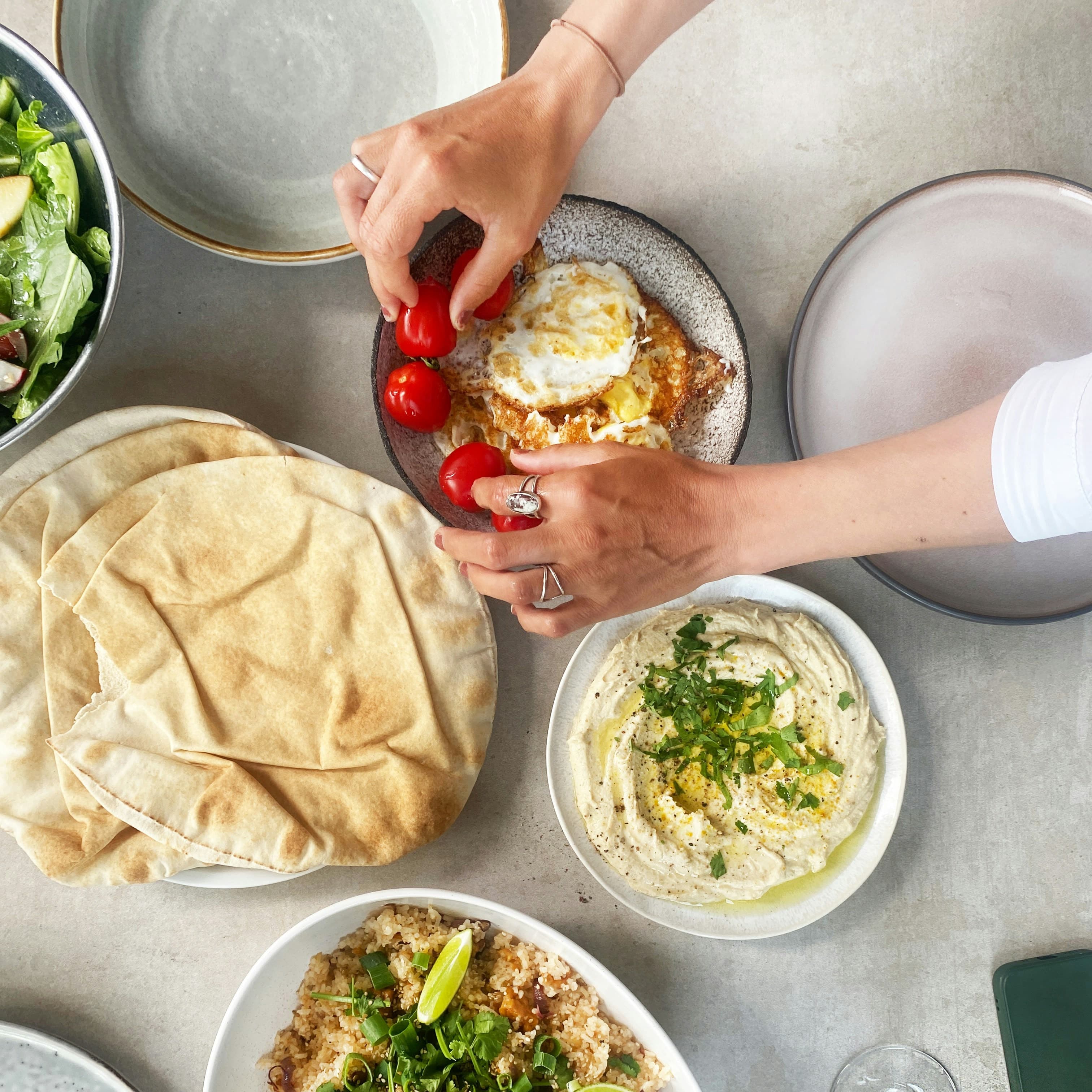 A photo of a table full of Mediterranean food taken from above. We can see a plate of pita, a bowl of hummus, a bowl with fried eggs and cherry tomatoes, a rice dish with limes and greens on top, and the corner of a green salad. We can also see several empty plates. A woman's hands reach out over the food and she is touching some of the cherry tomatoes as though arranging them on the plate.
